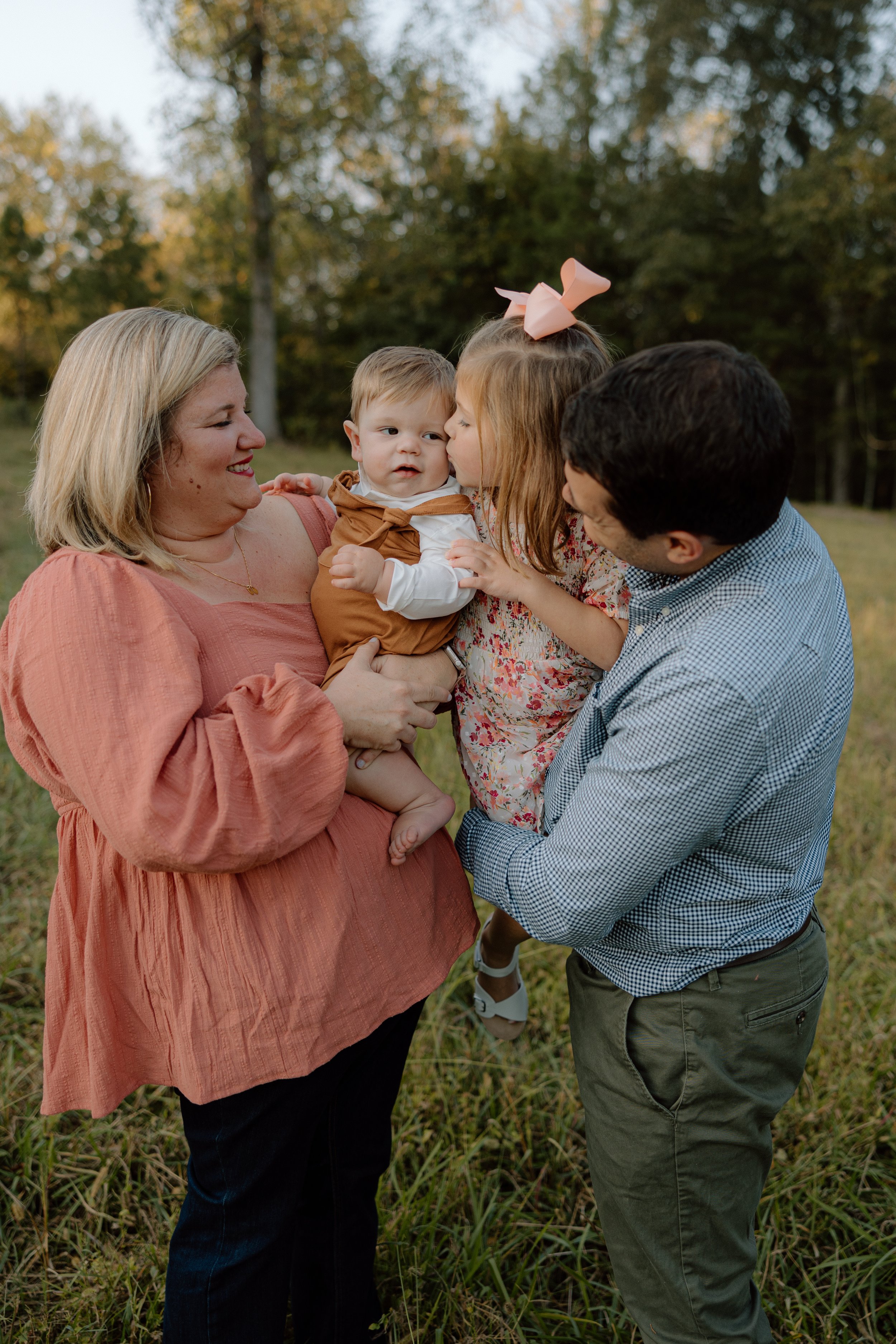 A family of five enjoying a moment outdoors in a grassy field with trees in the background. The mother is holding a young boy, while a girl kisses the boy on the cheek, and the father supports the girl.