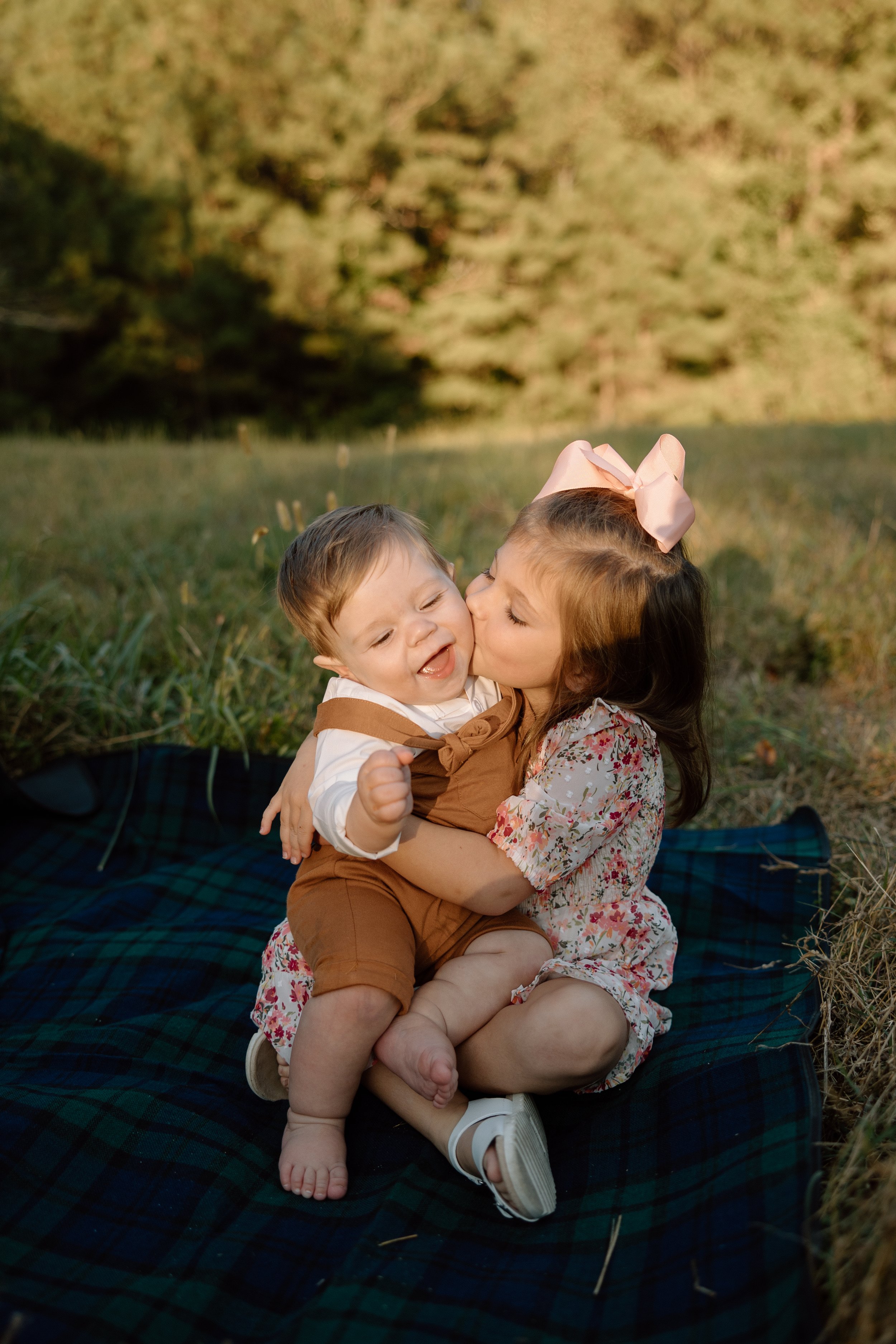 A young girl with a pink bow in her hair is hugging and kissing a young boy on the cheek as they sit on a blanket in a grassy field during sunset.