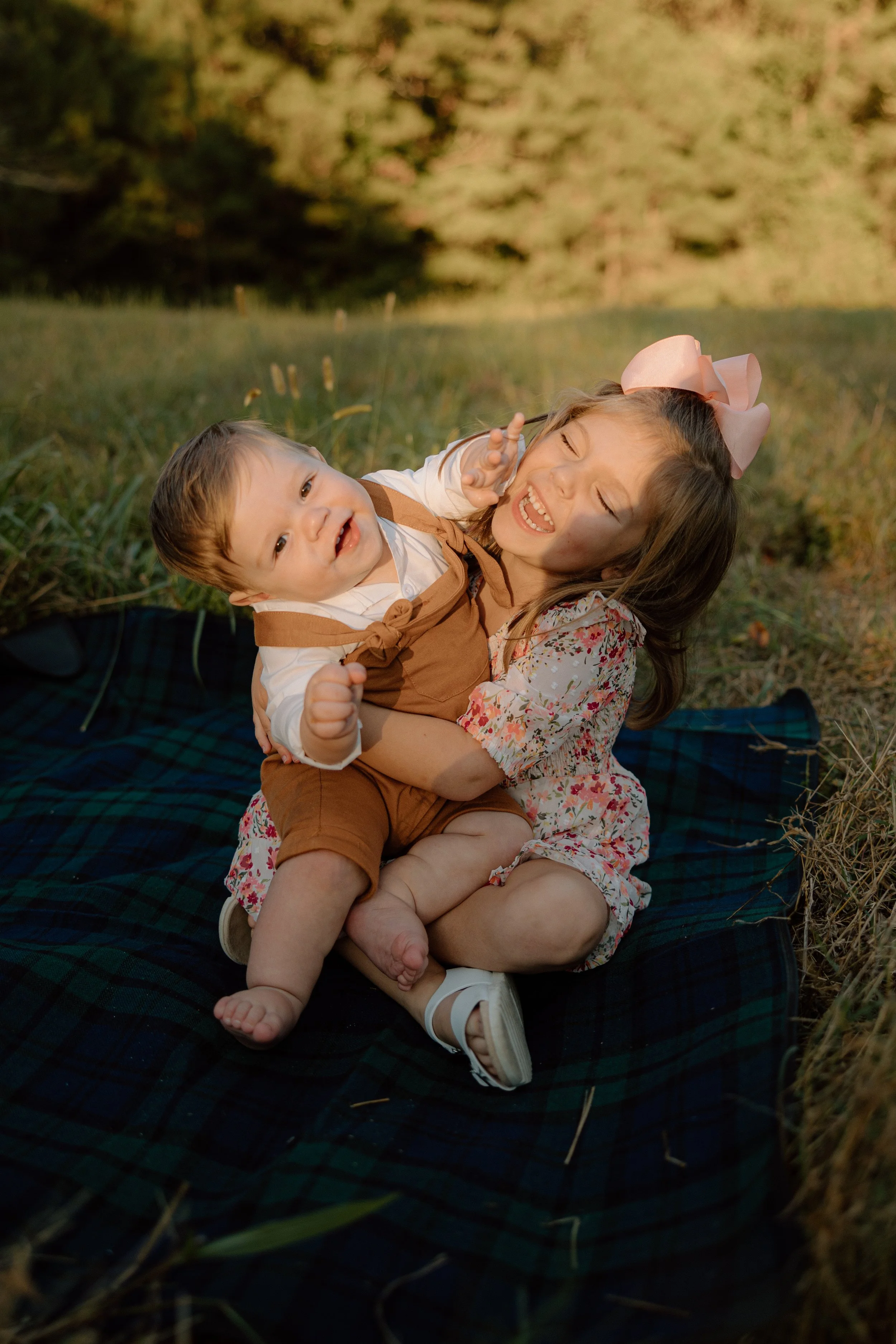 Young girl with a pink bow and a floral dress playing with a toddler in a field with a blanket at sunset.