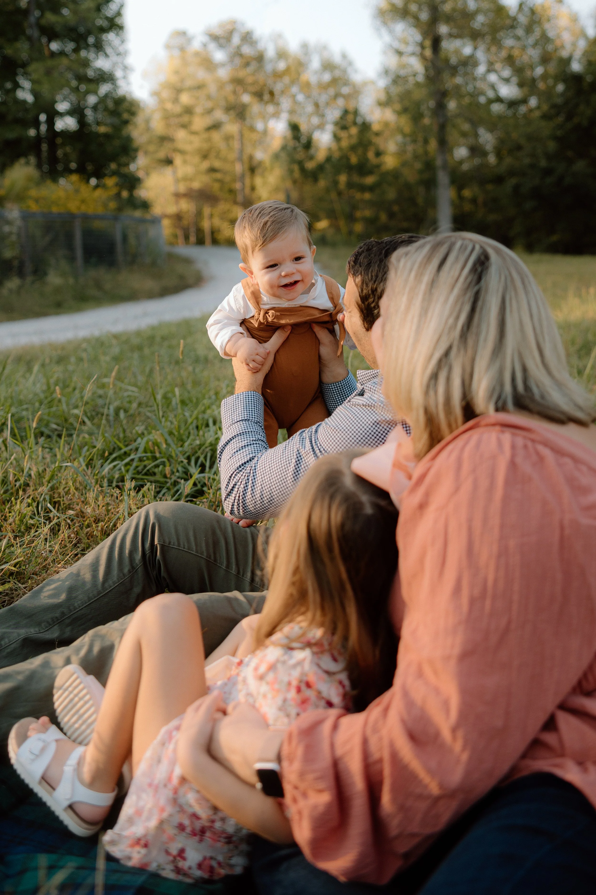 A family sitting on grass outdoors in a park or field with trees, enjoying a sunset. A man is holding a smiling toddler, and a woman with a girl are nearby.