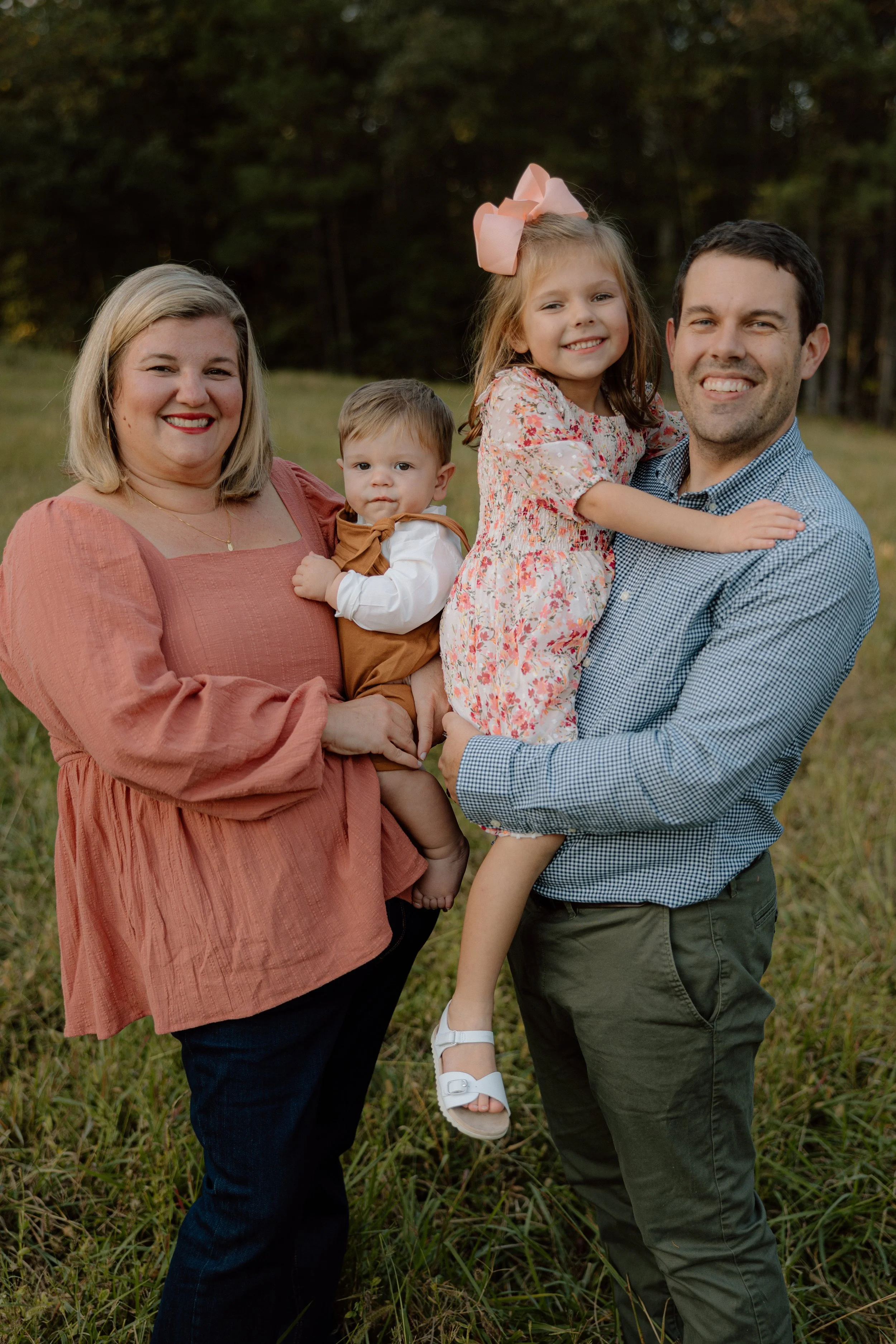Family of four in a grassy field, smiling at the camera, with a wooded background. A woman in a pink blouse holding a young boy, a man holding a young girl with a pink bow, and all appear happy and outdoors.