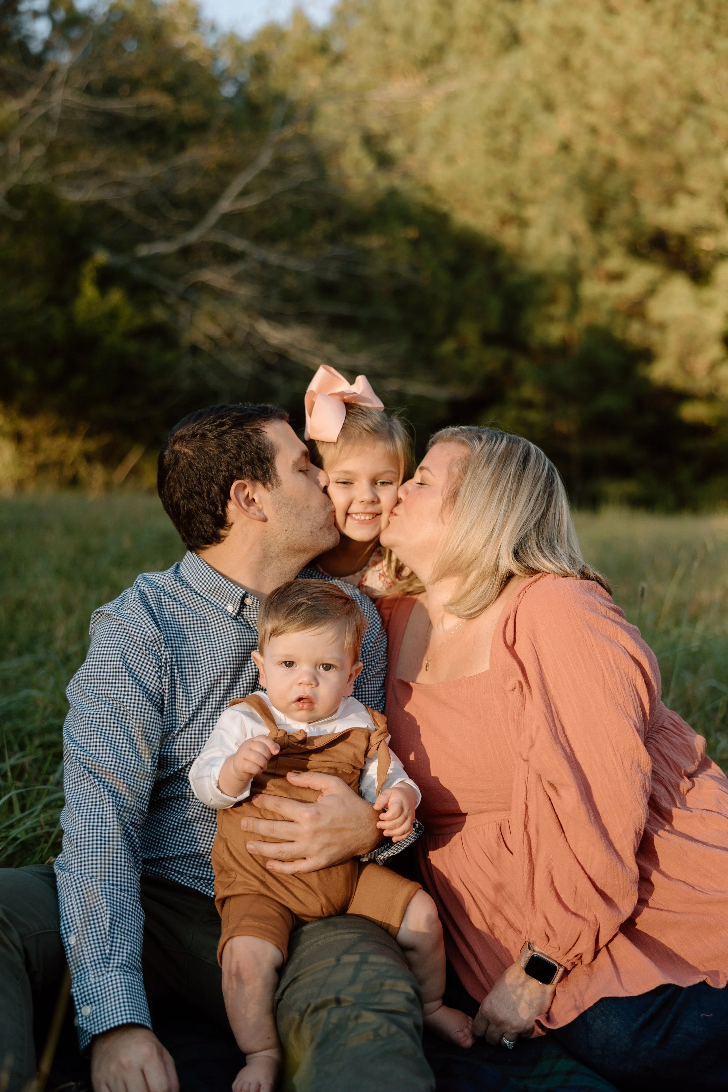A family of four sitting outdoors in a grassy field, with trees in the background. The father and mother are kissing their older daughter on the cheeks, while the younger son sits on the mother's lap, looking at the camera.