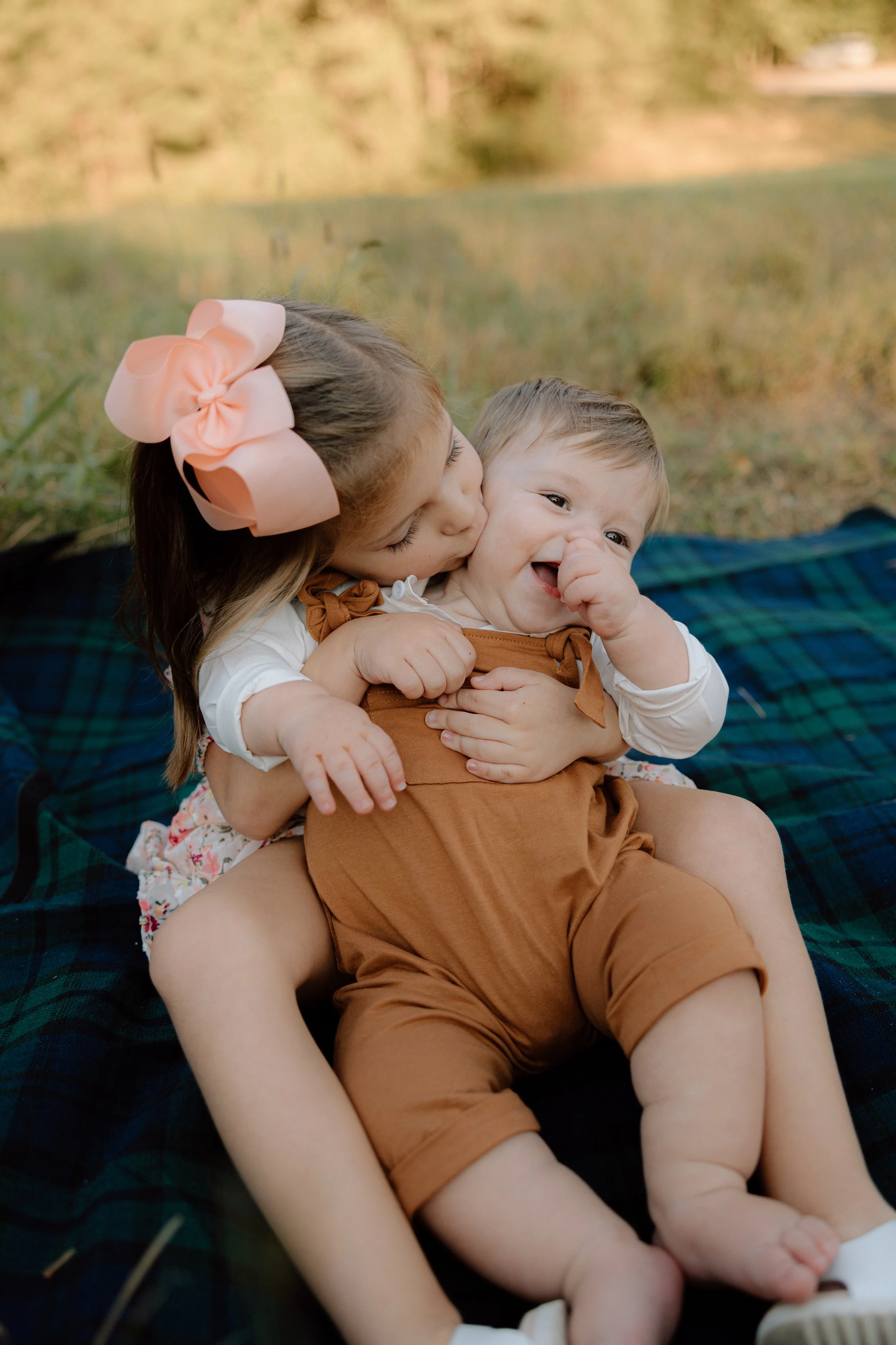 A young girl with a large pink bow in her hair is hugging and kissing a laughing baby boy on a blanket outdoors.