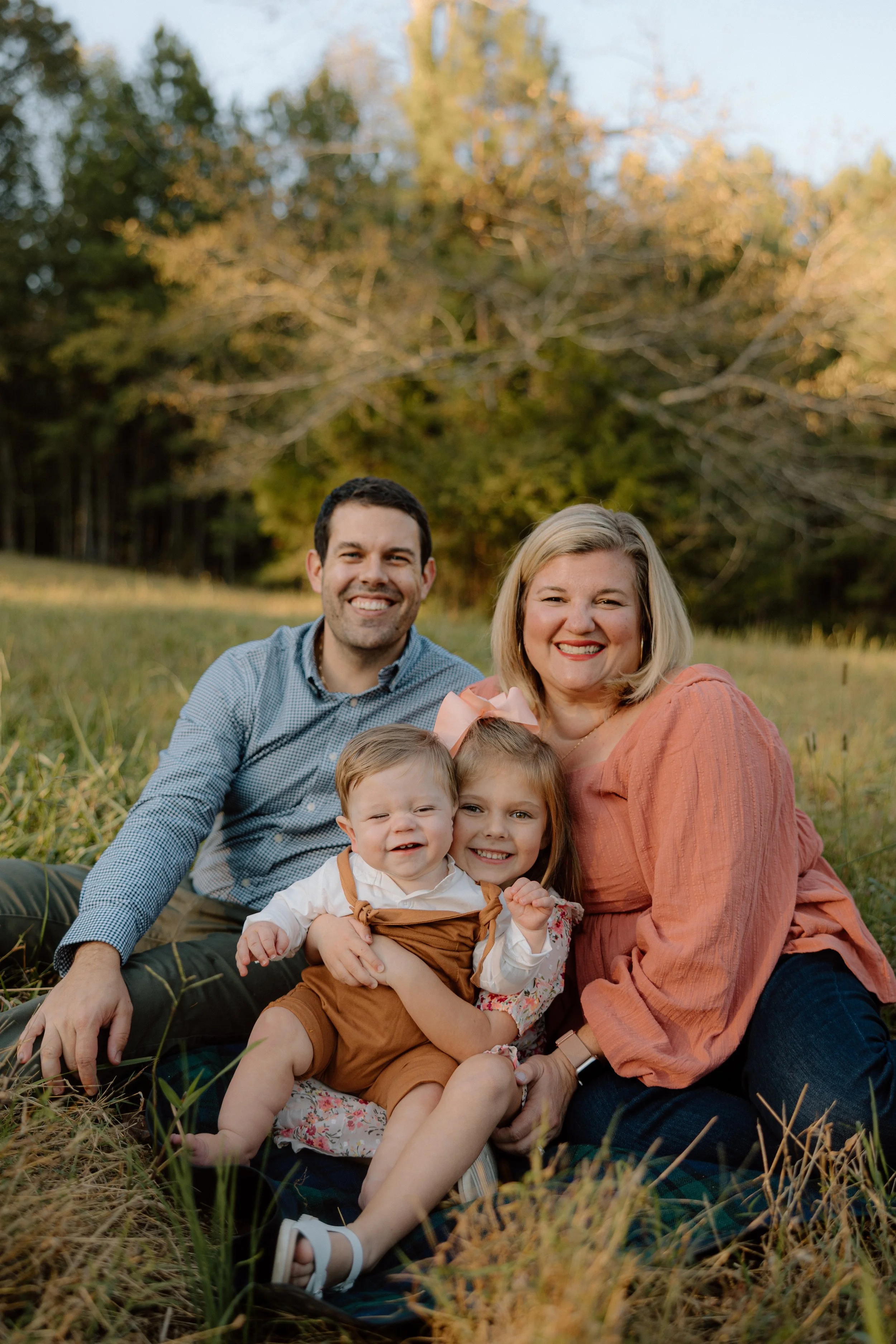 A family of four sitting on grass in a park during autumn, smiling at the camera.