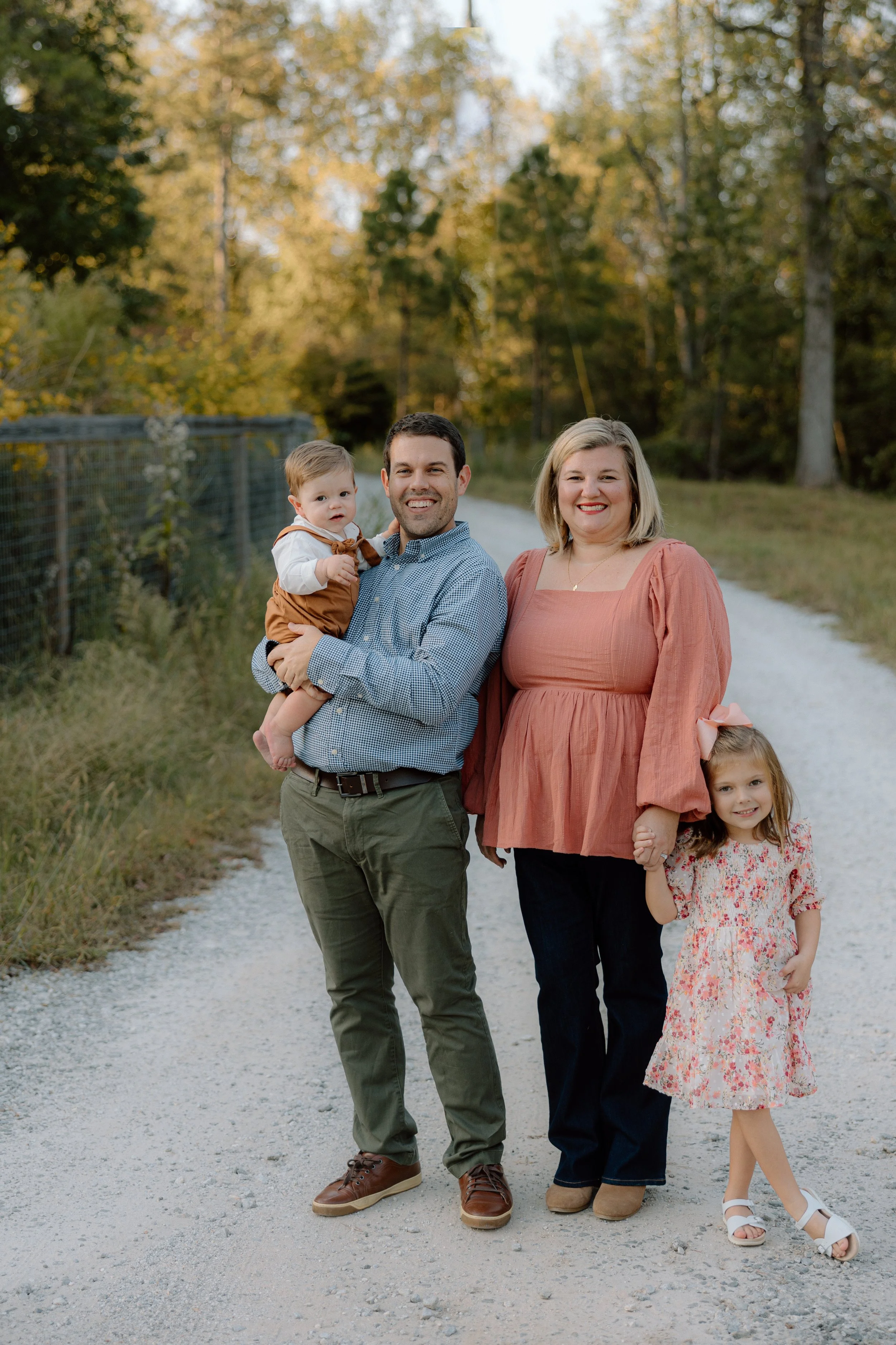 A family of four standing together outdoors on a gravel path, smiling at the camera during the daytime in a natural setting with trees in the background. The father is holding a young boy, and the mother is holding hands with a young girl.