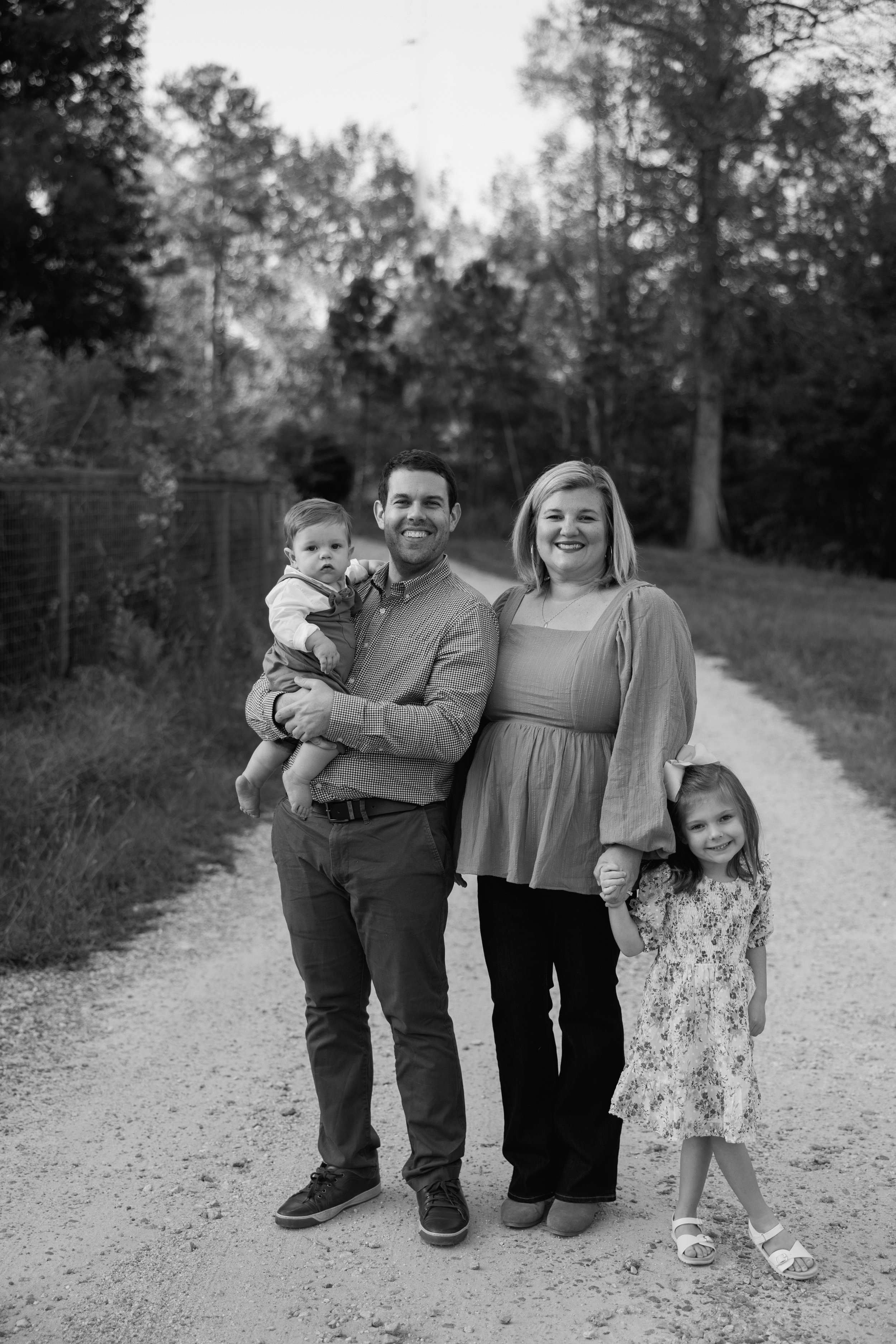 A black-and-white photo of a family of four standing on a dirt path outdoors, surrounded by trees. The father is holding a toddler boy, and the mother is holding the hand of a young girl. All are smiling.