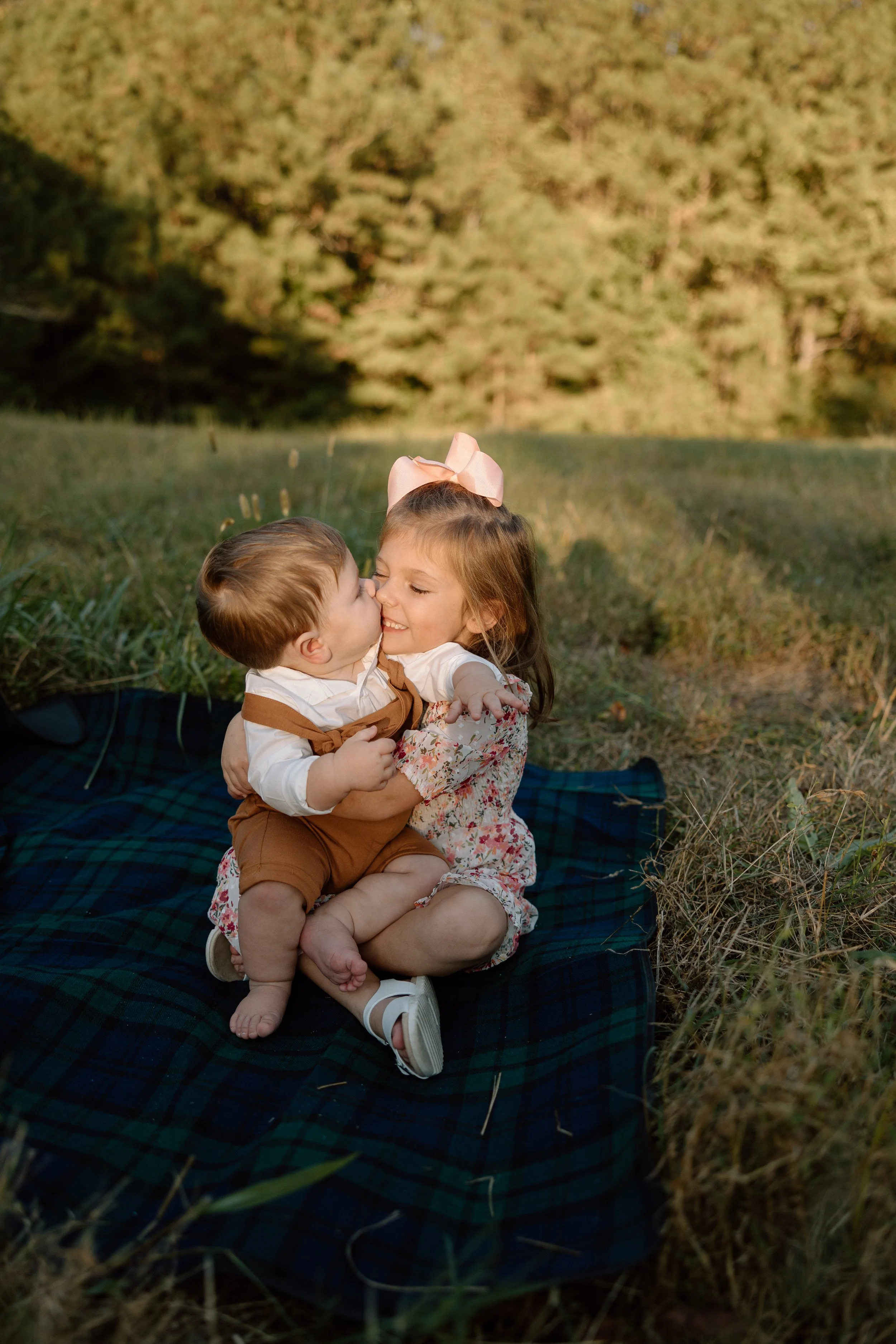Two young children, a girl and a boy, sitting on a blanket outdoors, sharing a joyful embrace and kiss in a grassy field with trees in the background.