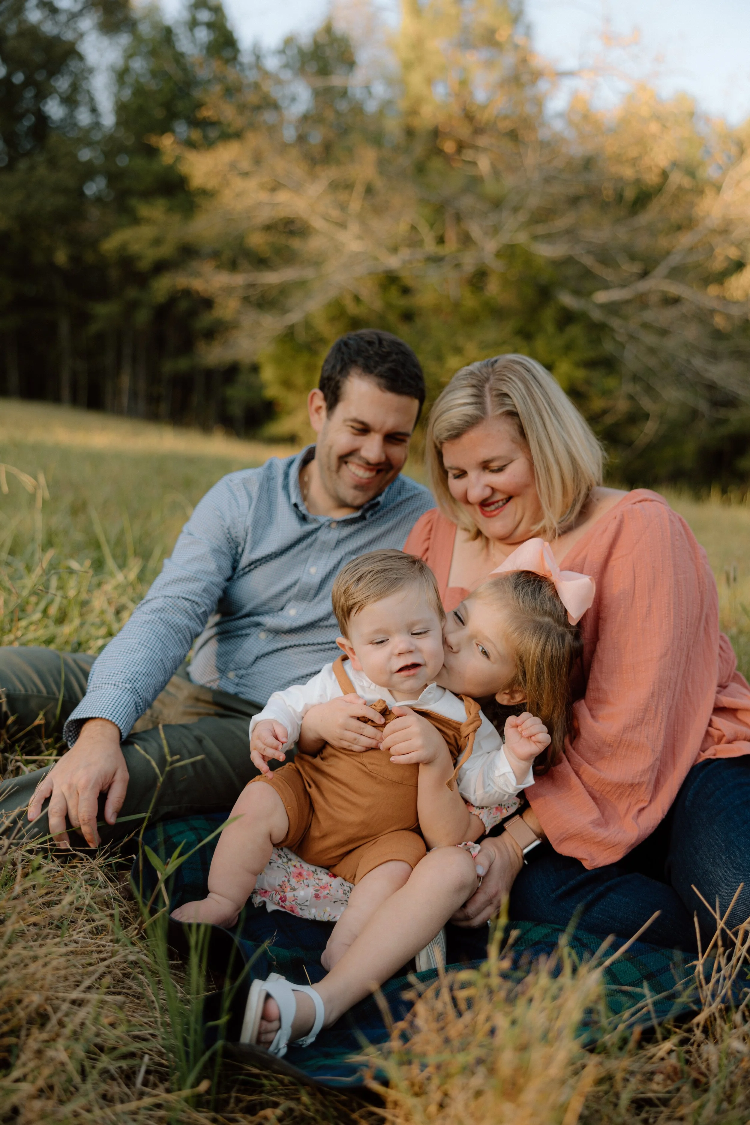 A happy family of four sitting on grass in a park or field during sunset, with trees and a blue sky in the background. The family includes a smiling man, a woman, a young girl, and a baby boy, with the girl kissing the baby on the cheek.