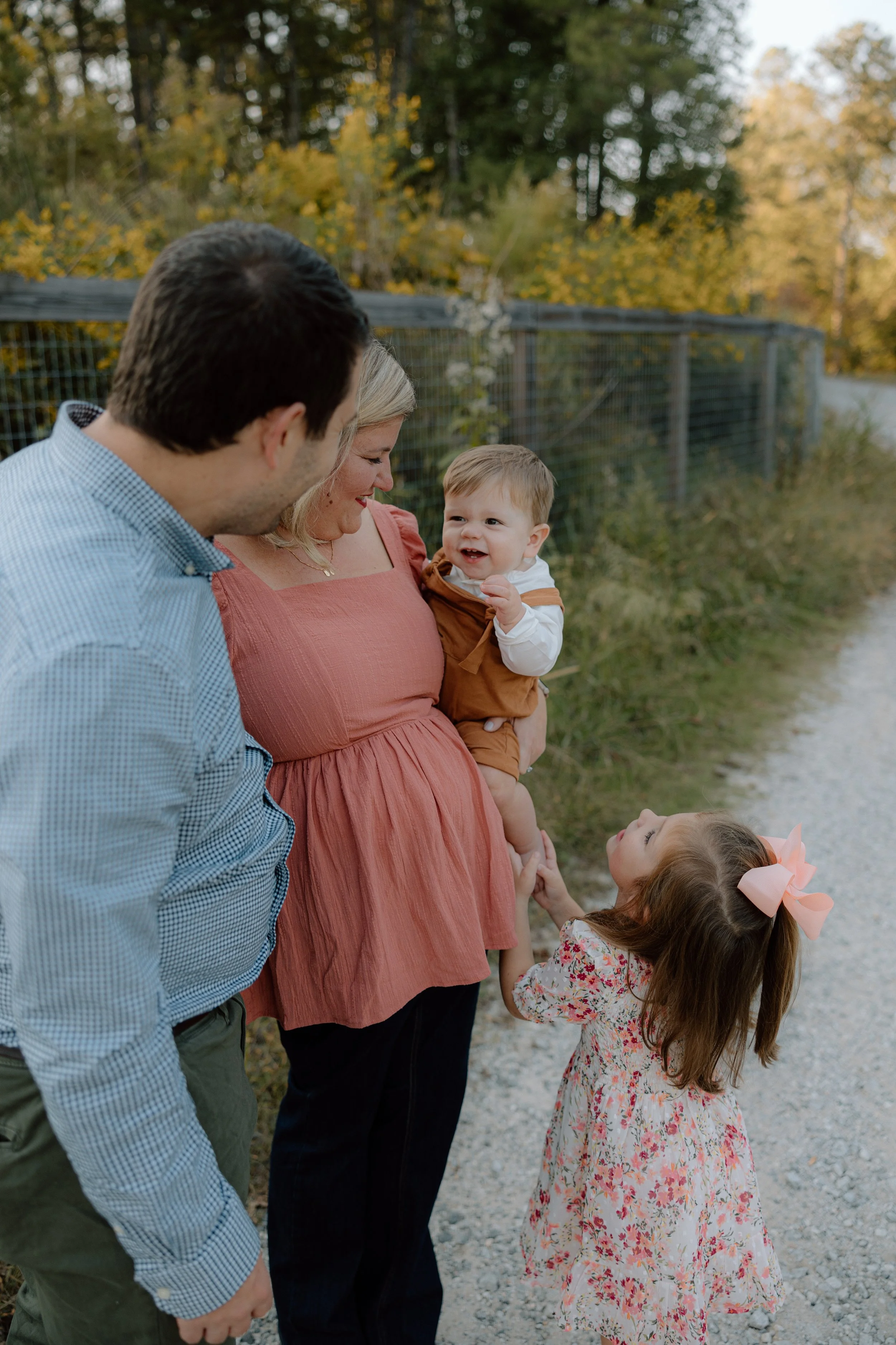 A family of four, parents and two children, smiling and sharing a moment outdoors on a gravel path with trees and a fence in the background during fall.