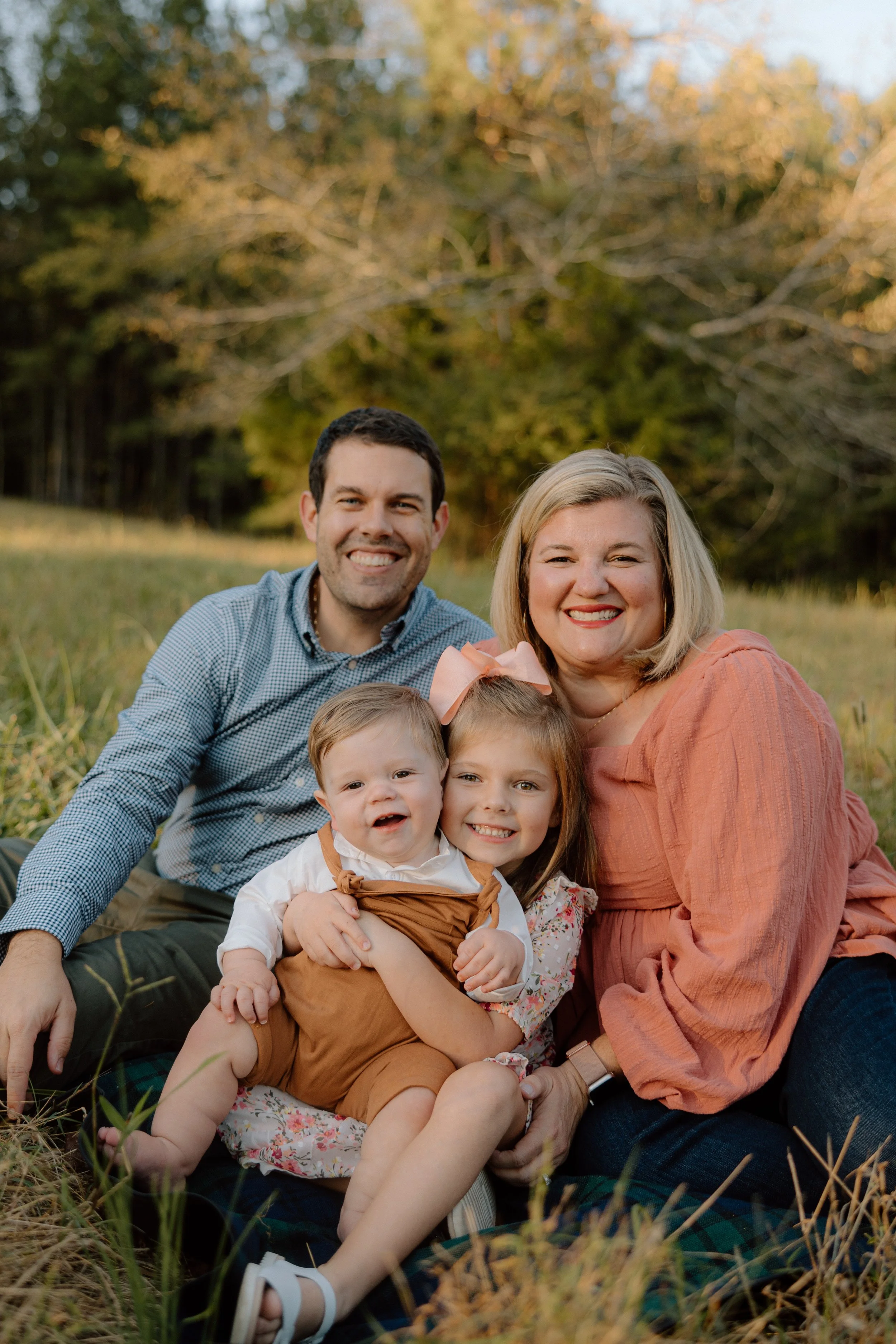 A happy family of four sitting outdoors on grass during fall, smiling at the camera with trees in the background.