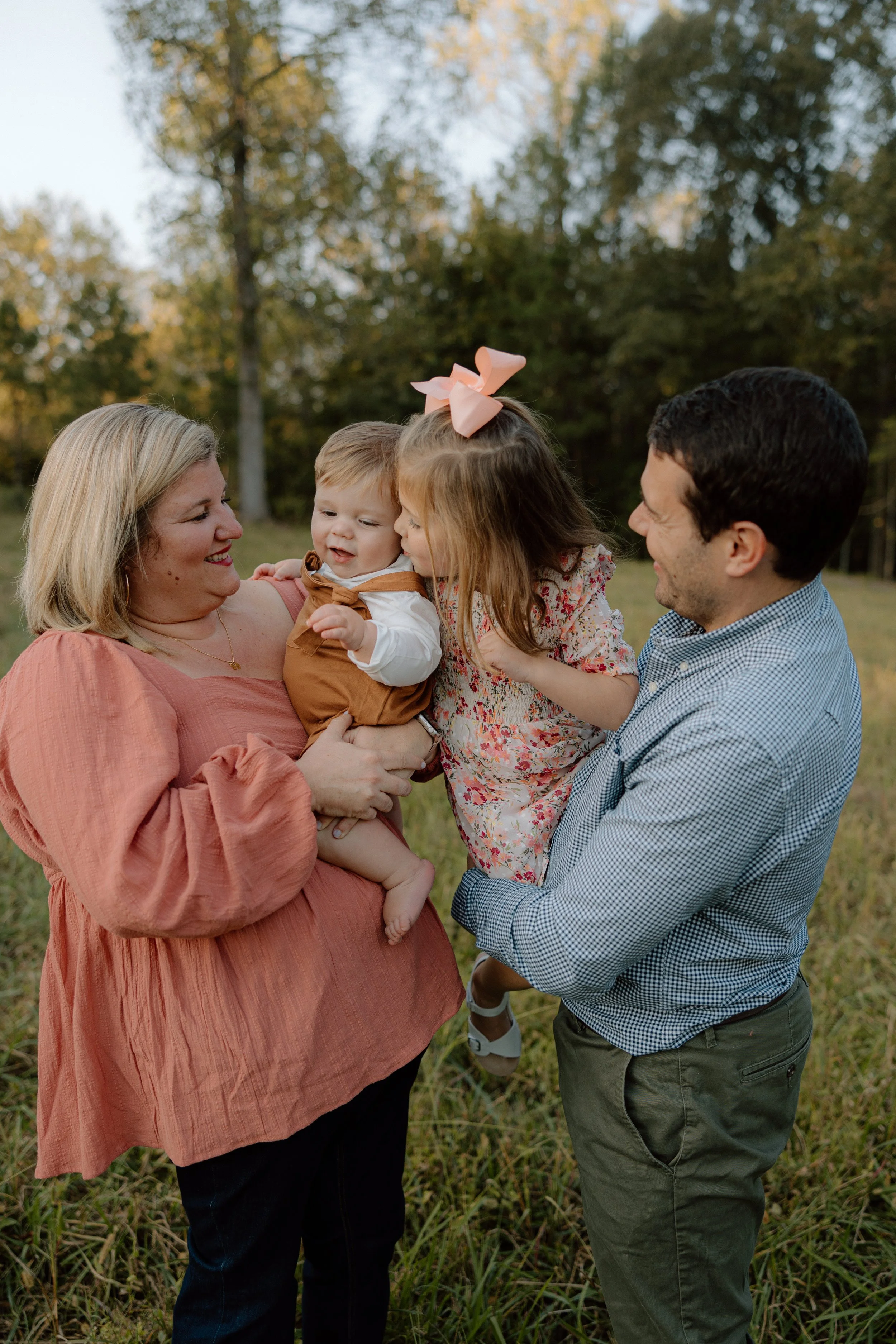 A family of four outdoors in a grassy field during golden hour, smiling and playing together. A woman with blonde hair wearing a pink blouse holds a toddler dressed in brown, while a man in a checkered shirt lifts a girl with a large pink bow in her 