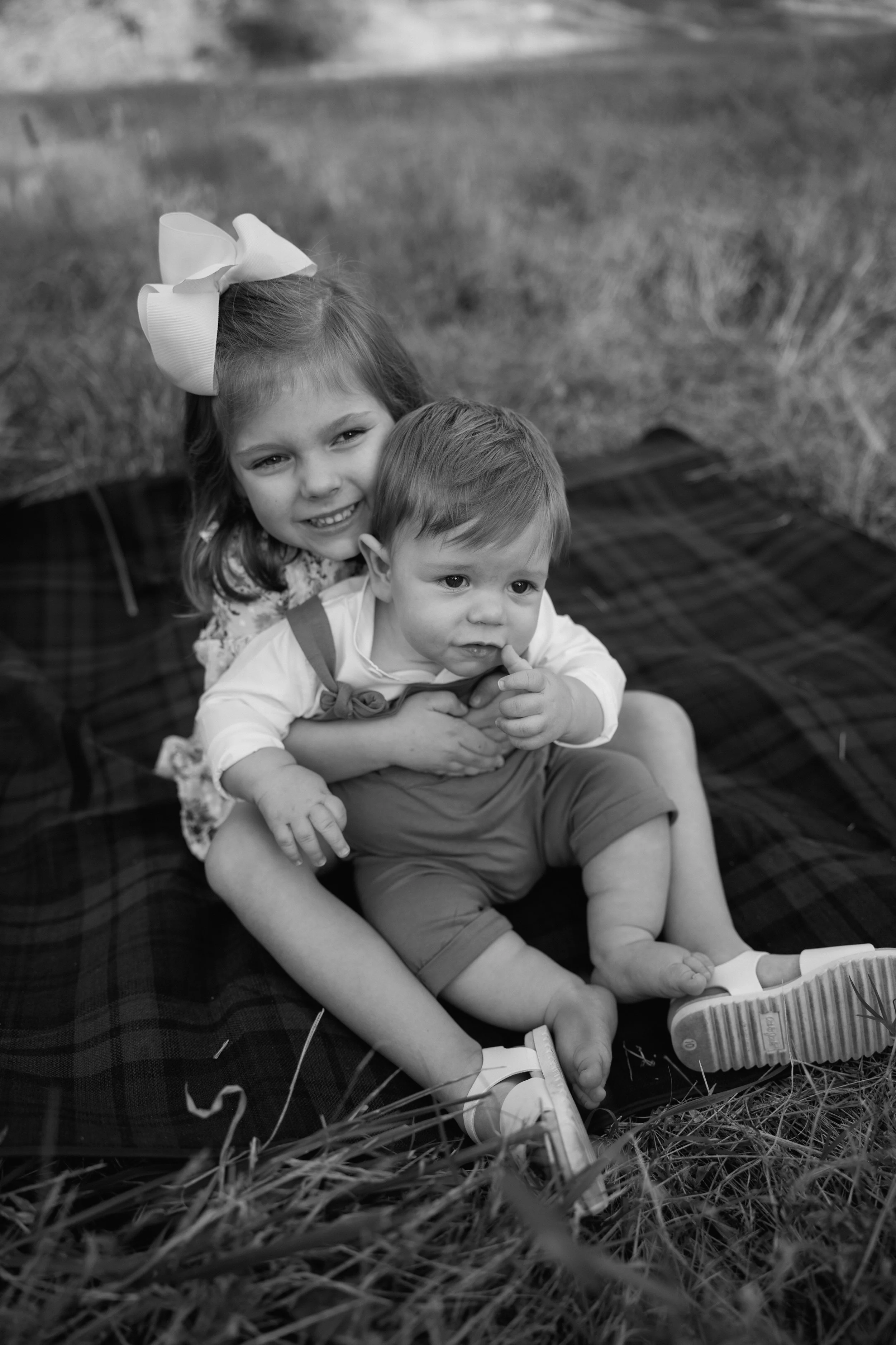 A young girl with a large bow in her hair holding a small boy on her lap, sitting on a blanket outdoors in a grassy field.