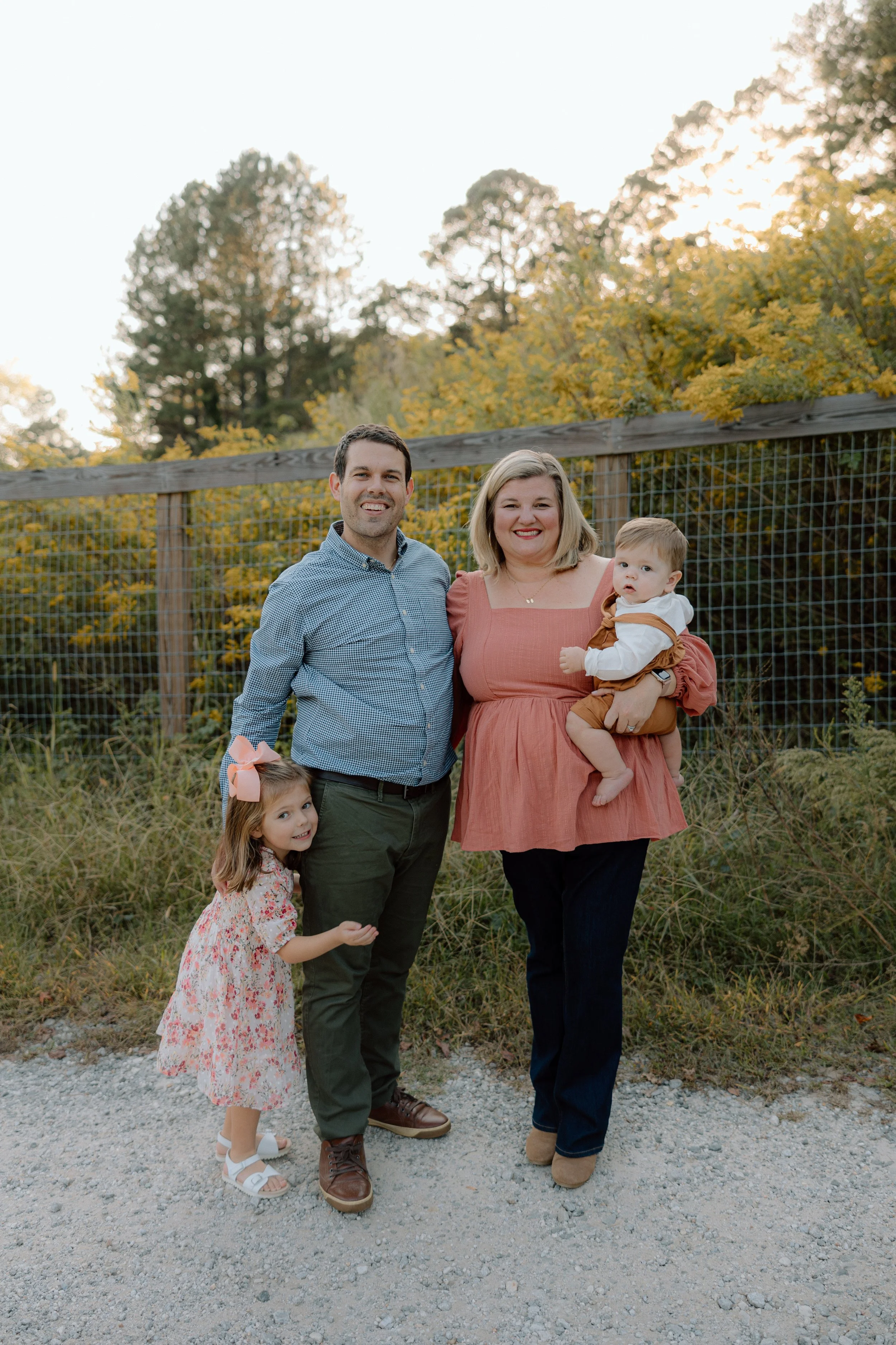 A family of four stands outdoors in front of a wooden fence and autumn trees, smiling at the camera. The mother holds a young child, and a daughter stands beside her father.