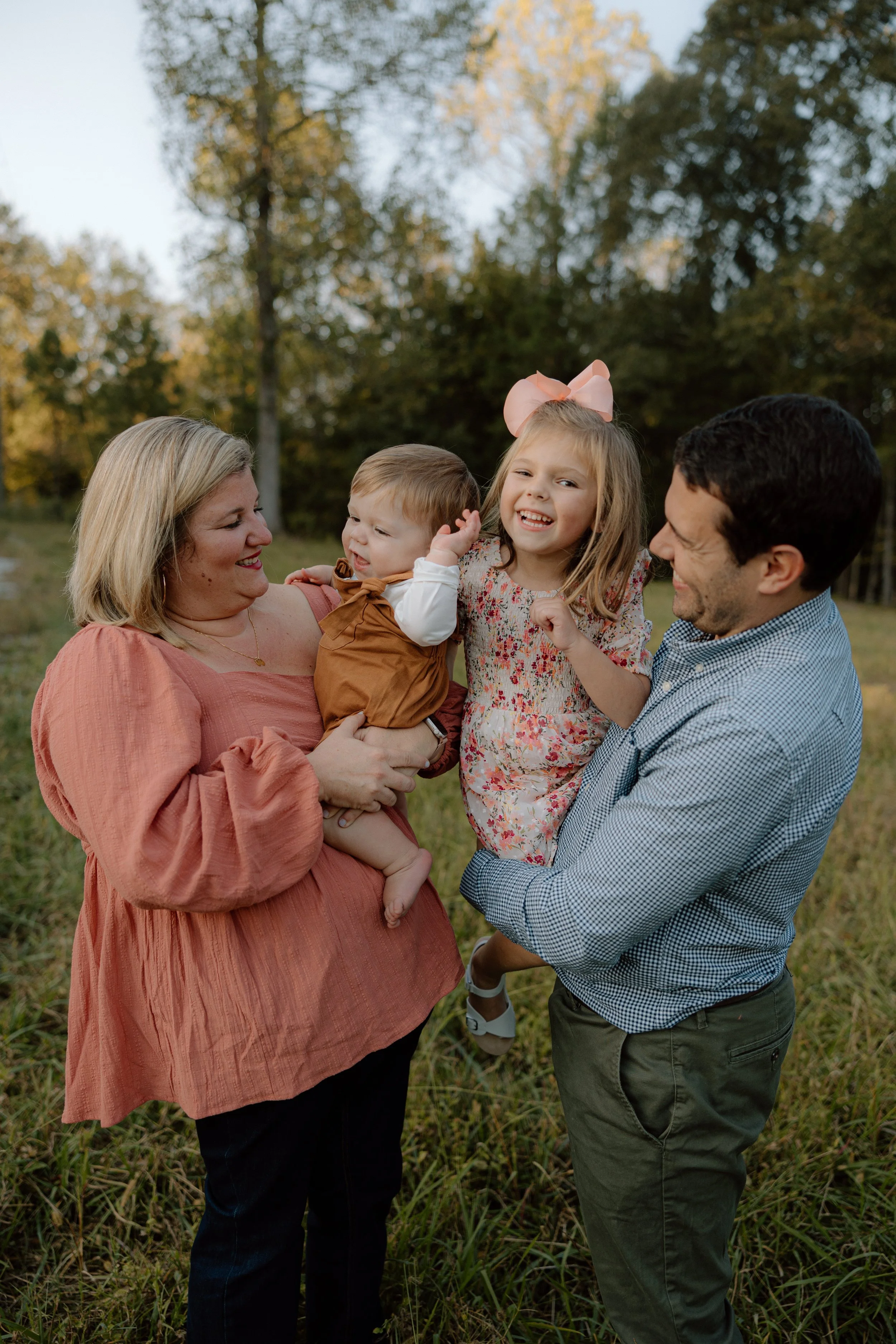 A family of four outdoors in a grassy area with trees in the background, smiling and playing together. The grandmother is holding a baby boy, and a man is holding a young girl with a pink bow in her hair.