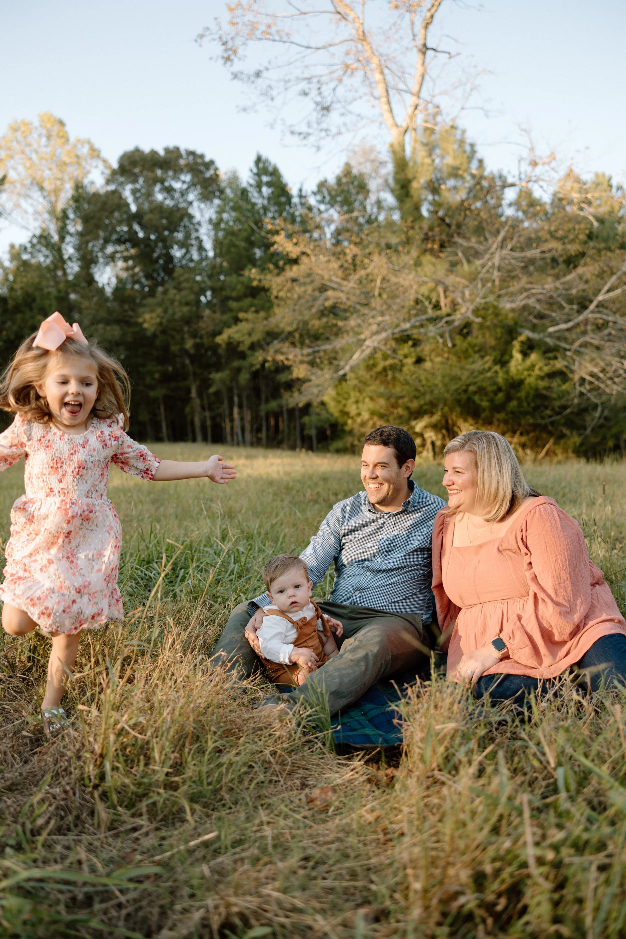 A family of four enjoying a picnic in a grassy field with trees in the background during autumn. The young girl is running playfully toward the seated parents and baby.