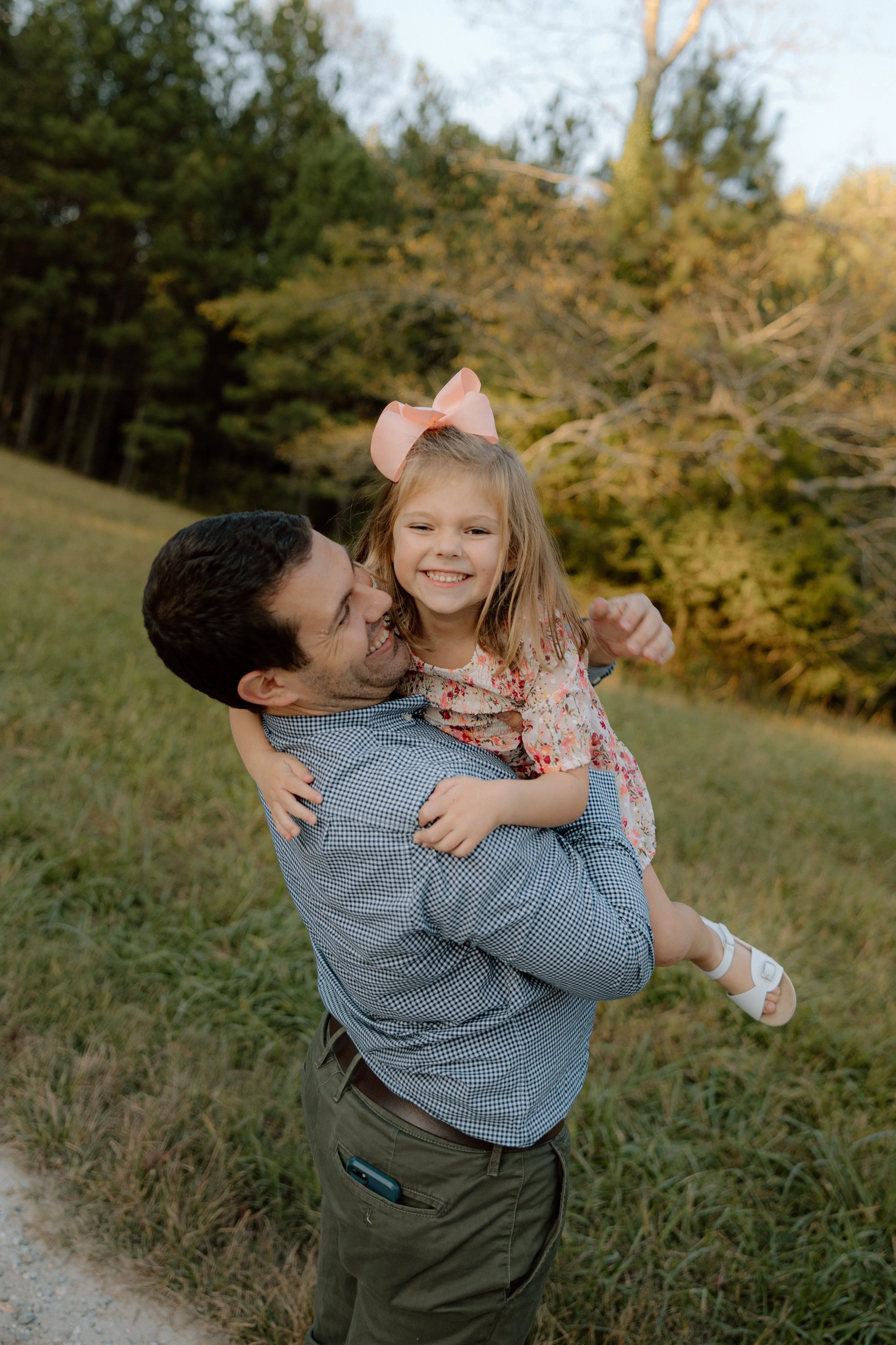 A man lifting a young girl with blonde hair and a pink bow, smiling and enjoying an outdoor moment in a grassy area with trees in the background.
