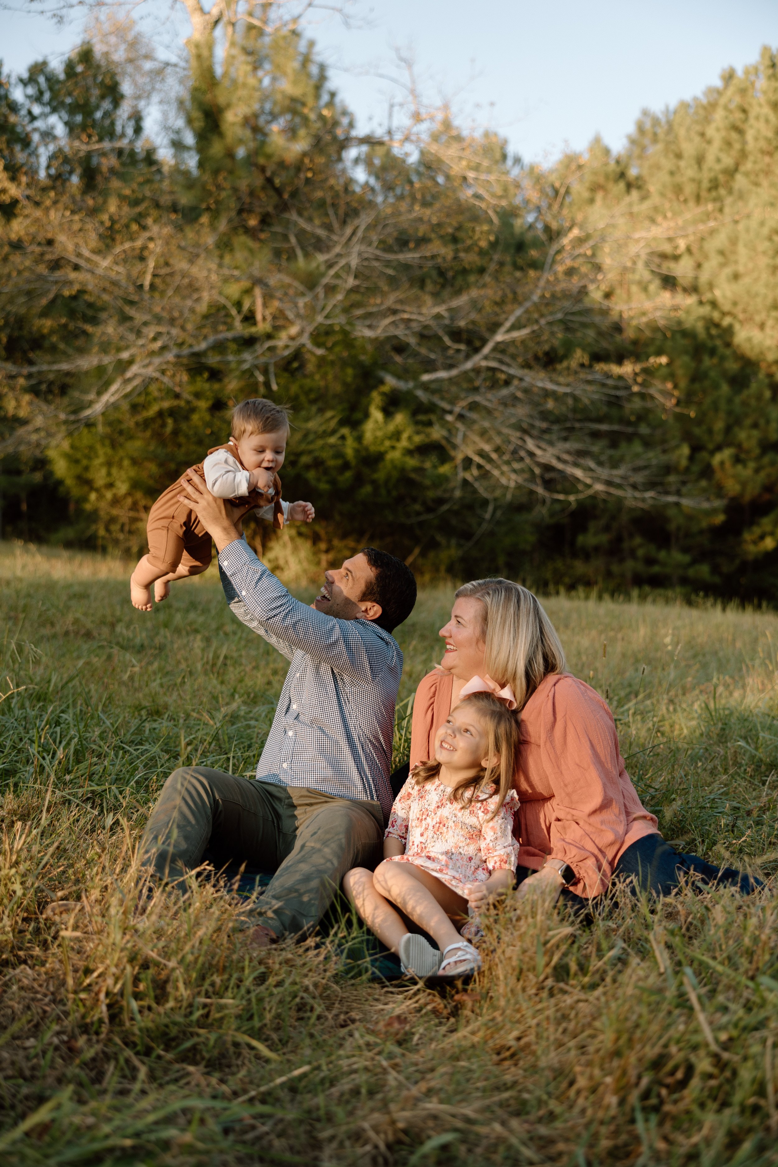A family of four sitting on the grass in a park during sunset, with a father lifting a baby boy into the air, a mother and young girl watching and smiling.