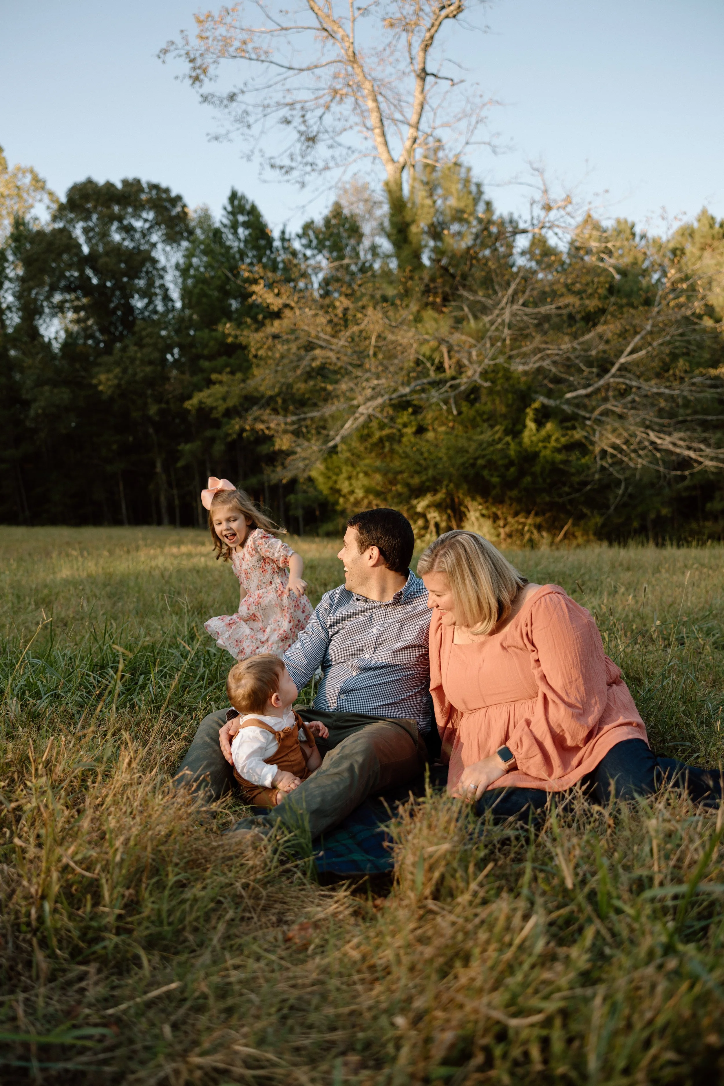 A family of four enjoying a picnic in a grassy field during late afternoon. The family includes a man, woman, young girl, and a baby. The young girl is running happily, and the parents are sitting on a blanket with the baby. The background shows tree
