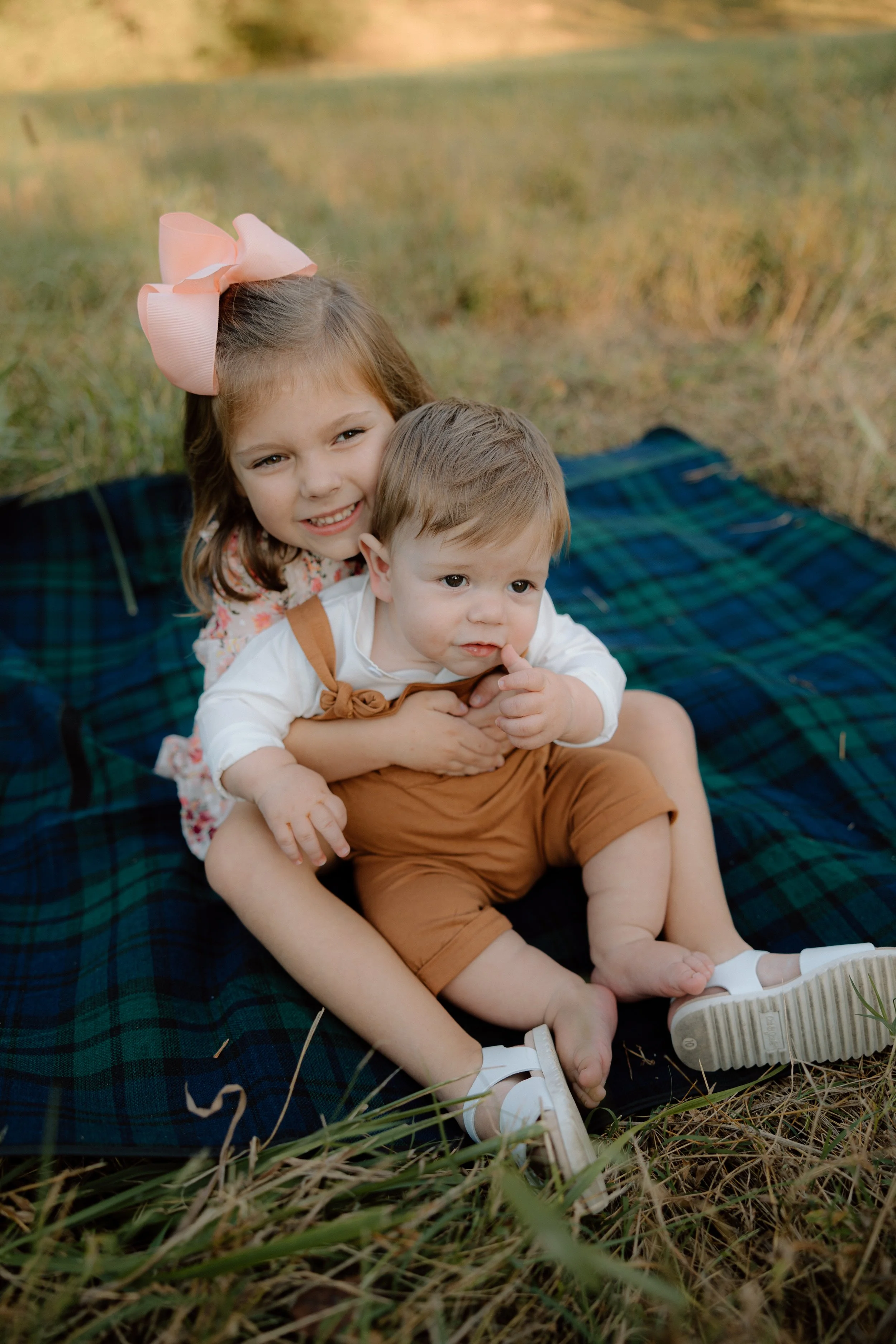 A young girl with a pink bow in her hair hugging a toddler boy while sitting on a plaid blanket outdoors.