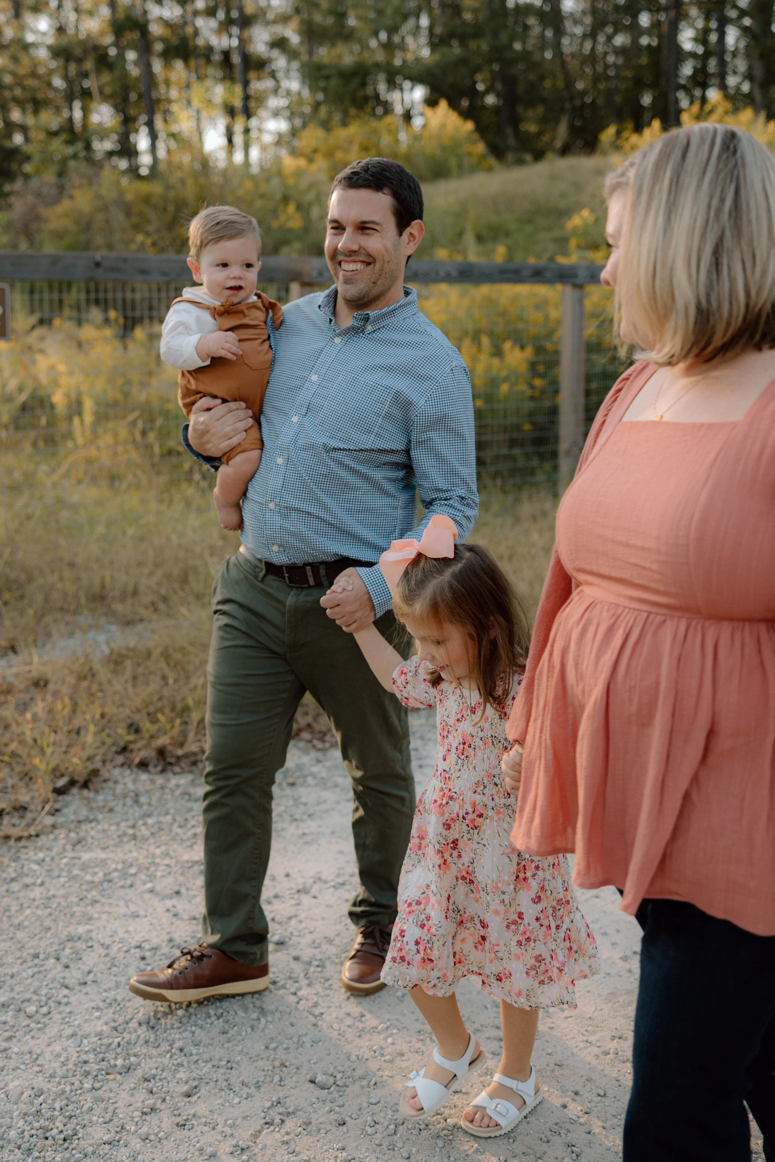 A family outdoors on a gravel path during fall, with three adults and two children, engaged in a joyful moment, surrounded by trees with autumn leaves.