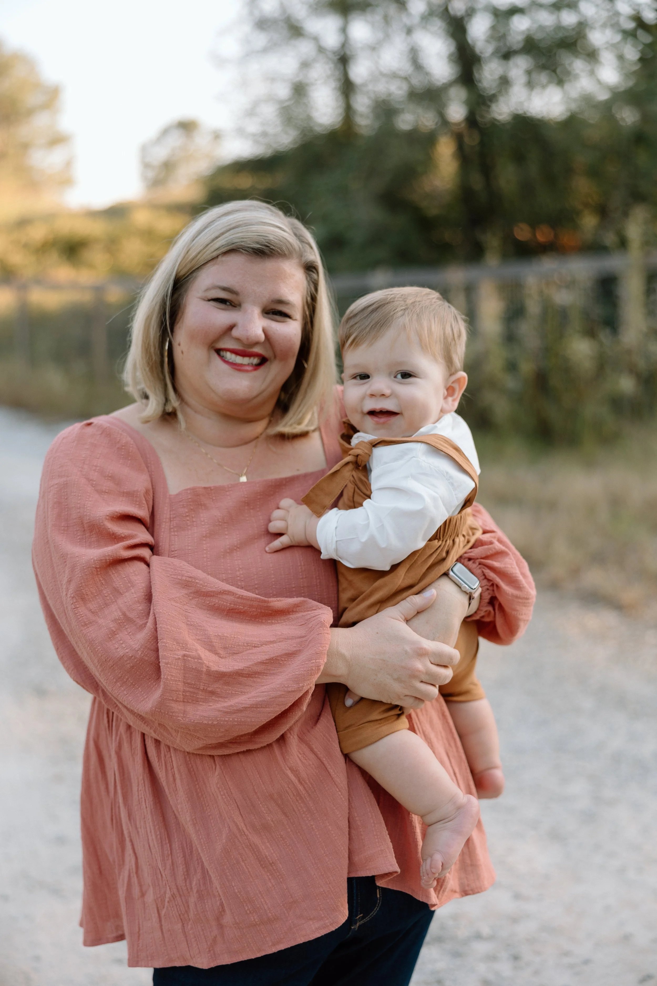 A smiling woman holding a young child outdoors near a fence with trees in the background.