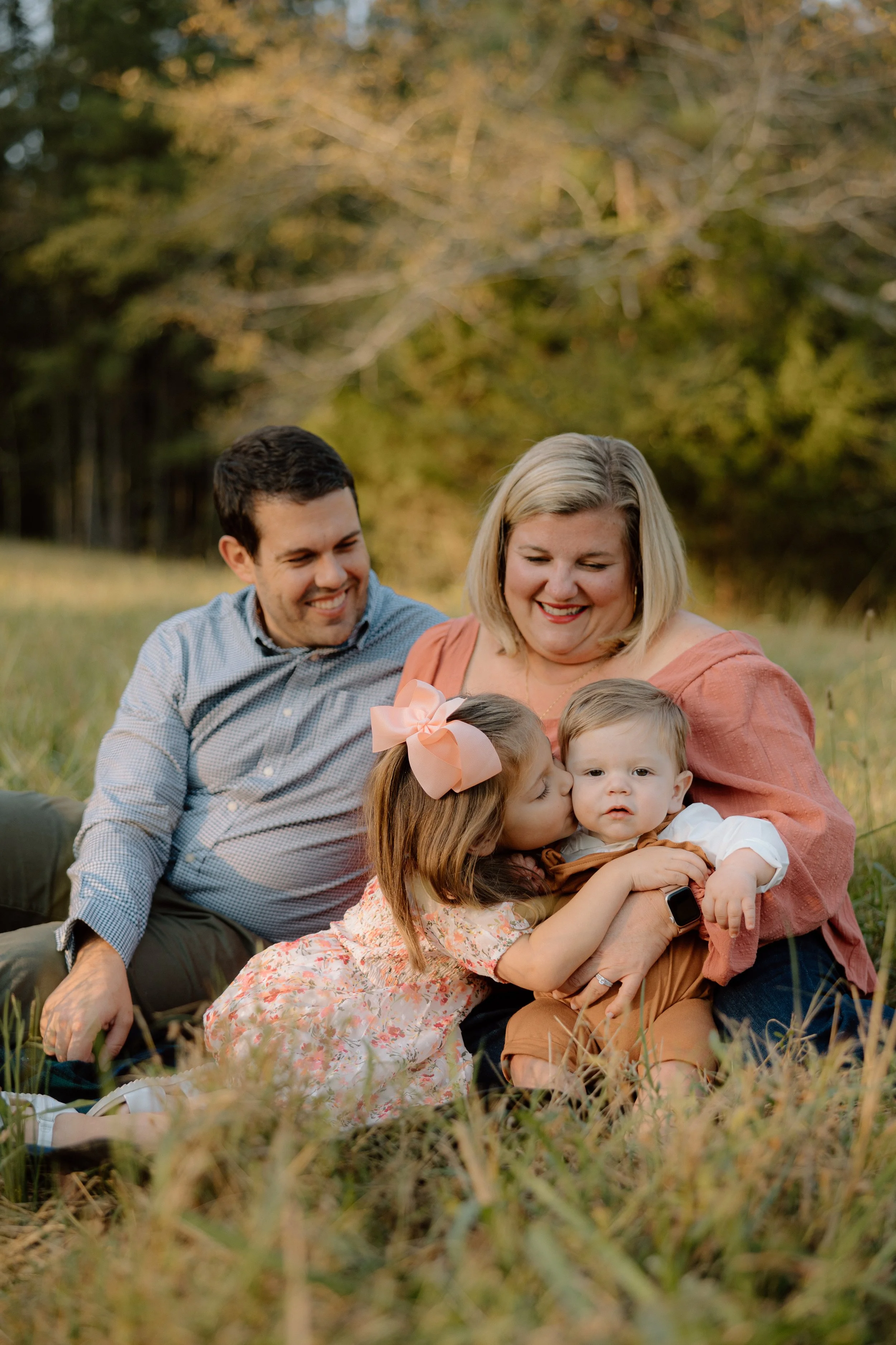 A family of four sitting on the grass in an outdoor setting with trees in the background. The mother is holding a young boy, and a girl is giving him a kiss on the cheek. The father is sitting beside them, smiling.