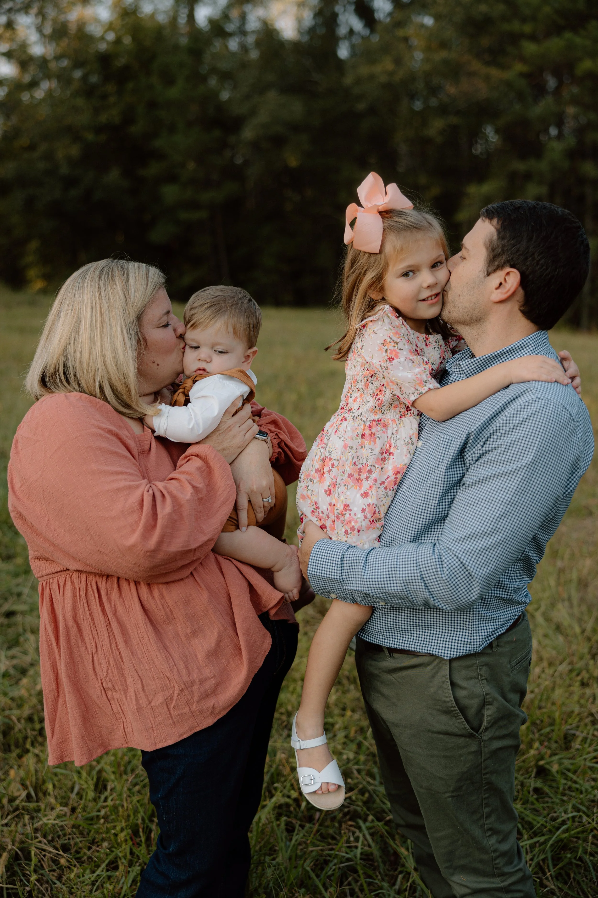 A family of four outdoors, with a mother holding a toddler boy, and a father holding a young girl who is kissing him on the lips. The background shows trees and grass.