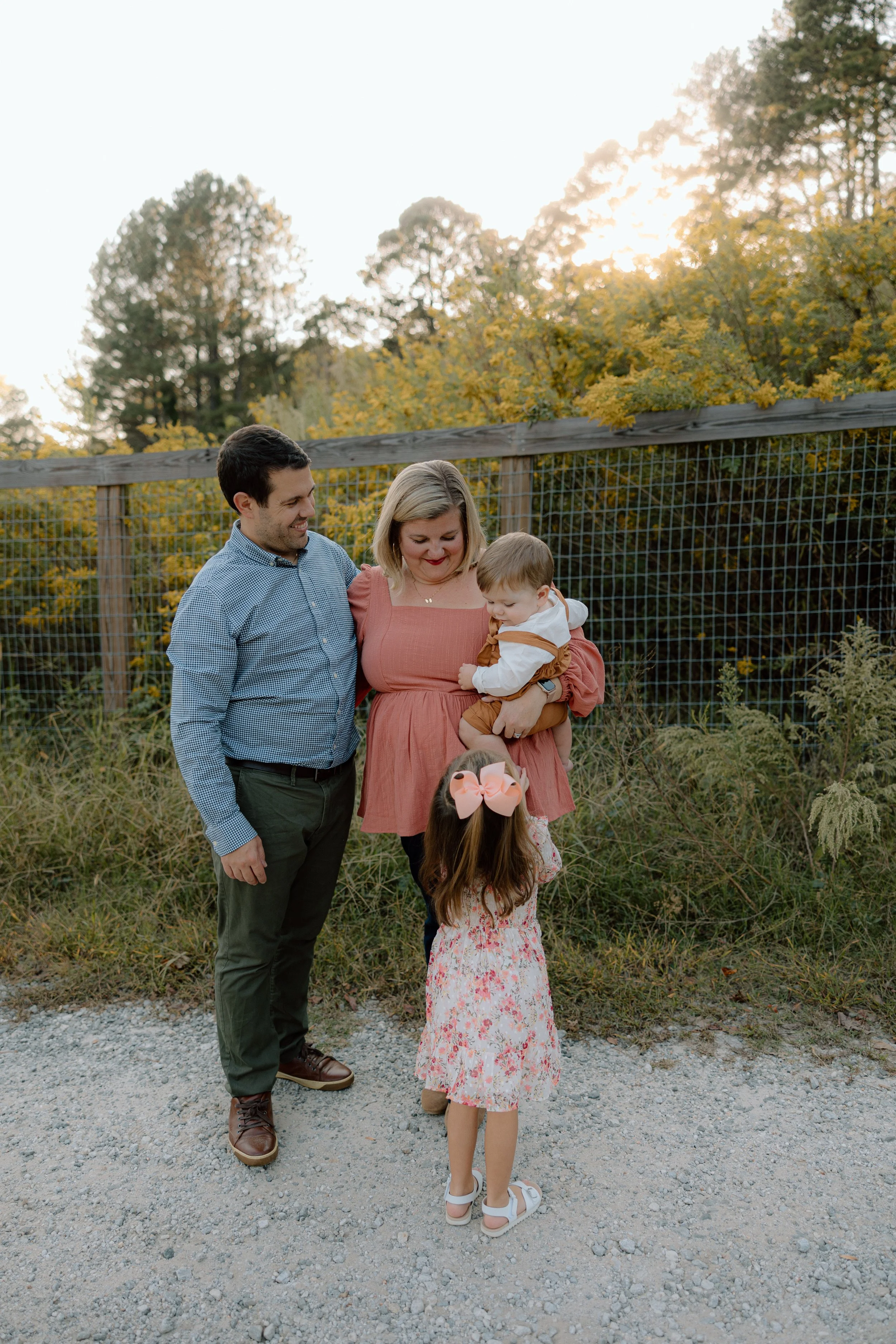 A family of four standing outdoors on a gravel path at sunset, with a wooden fence and trees in the background. The mother is holding a young boy, and a young girl with a large pink bow is standing in front of them, facing the mother.