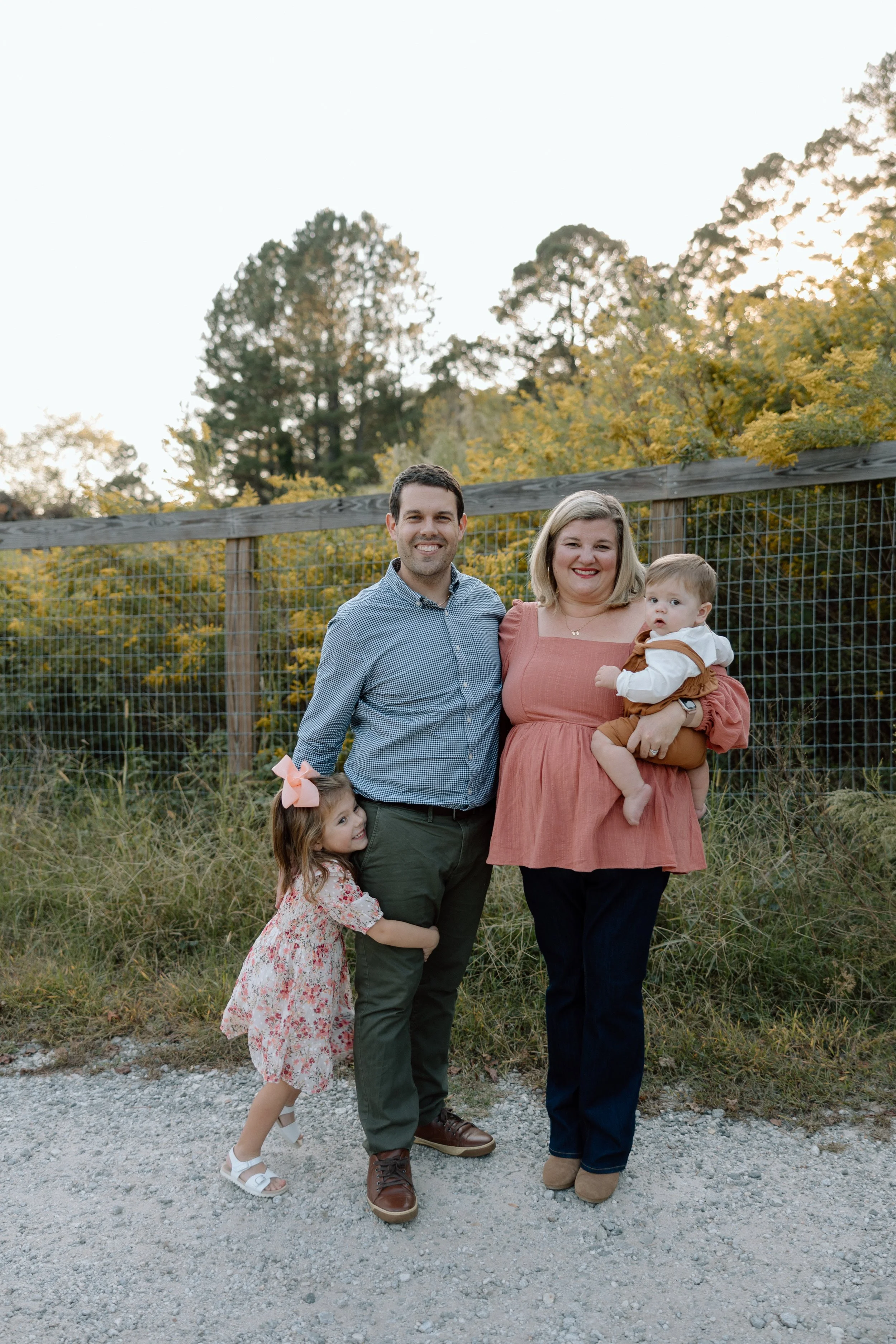 Family of four standing outdoors near a wooden fence, with trees and sunlight in the background, smiling at the camera.