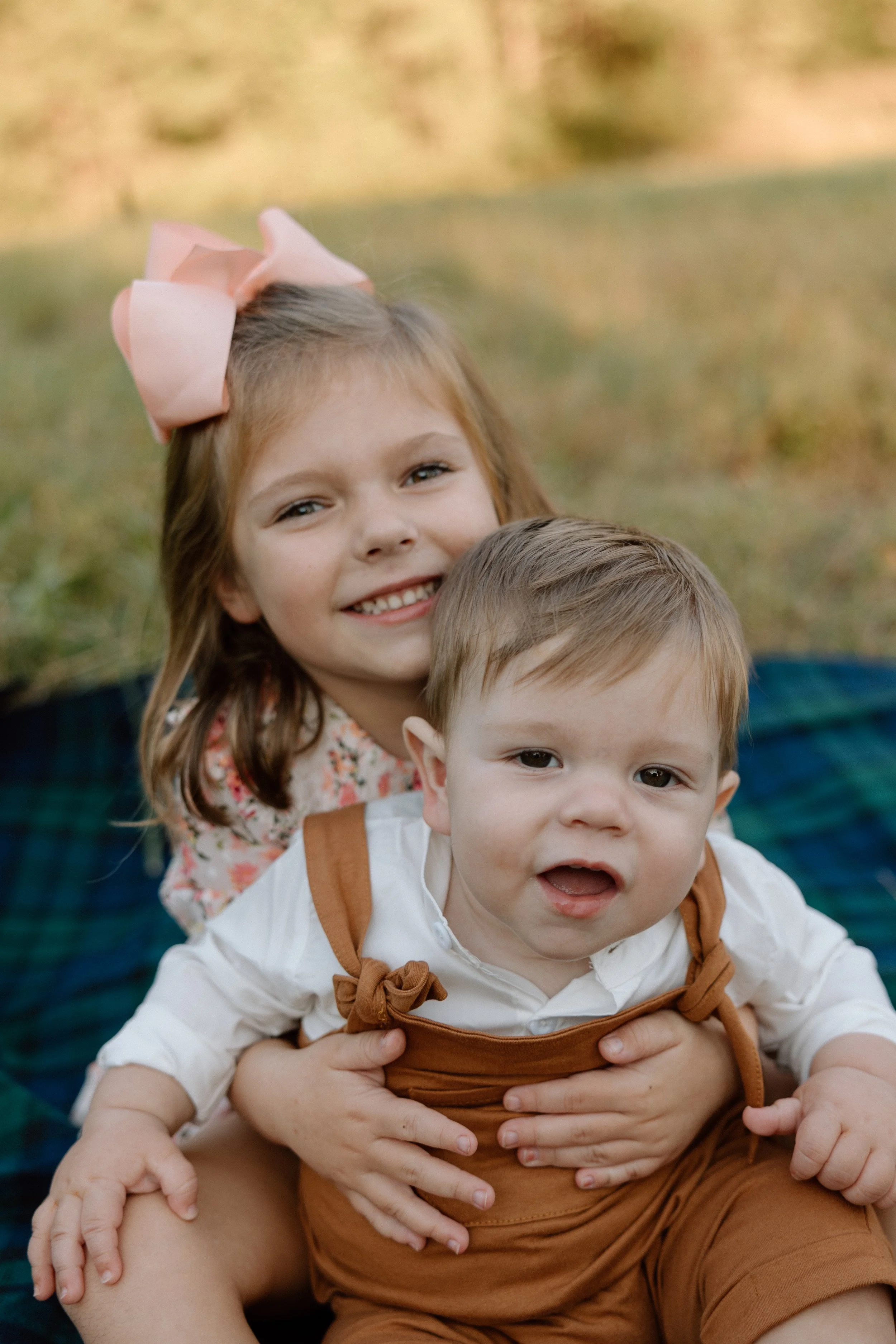 A young girl with a pink bow in her hair hugging a baby boy with brown hair while sitting outdoors.
