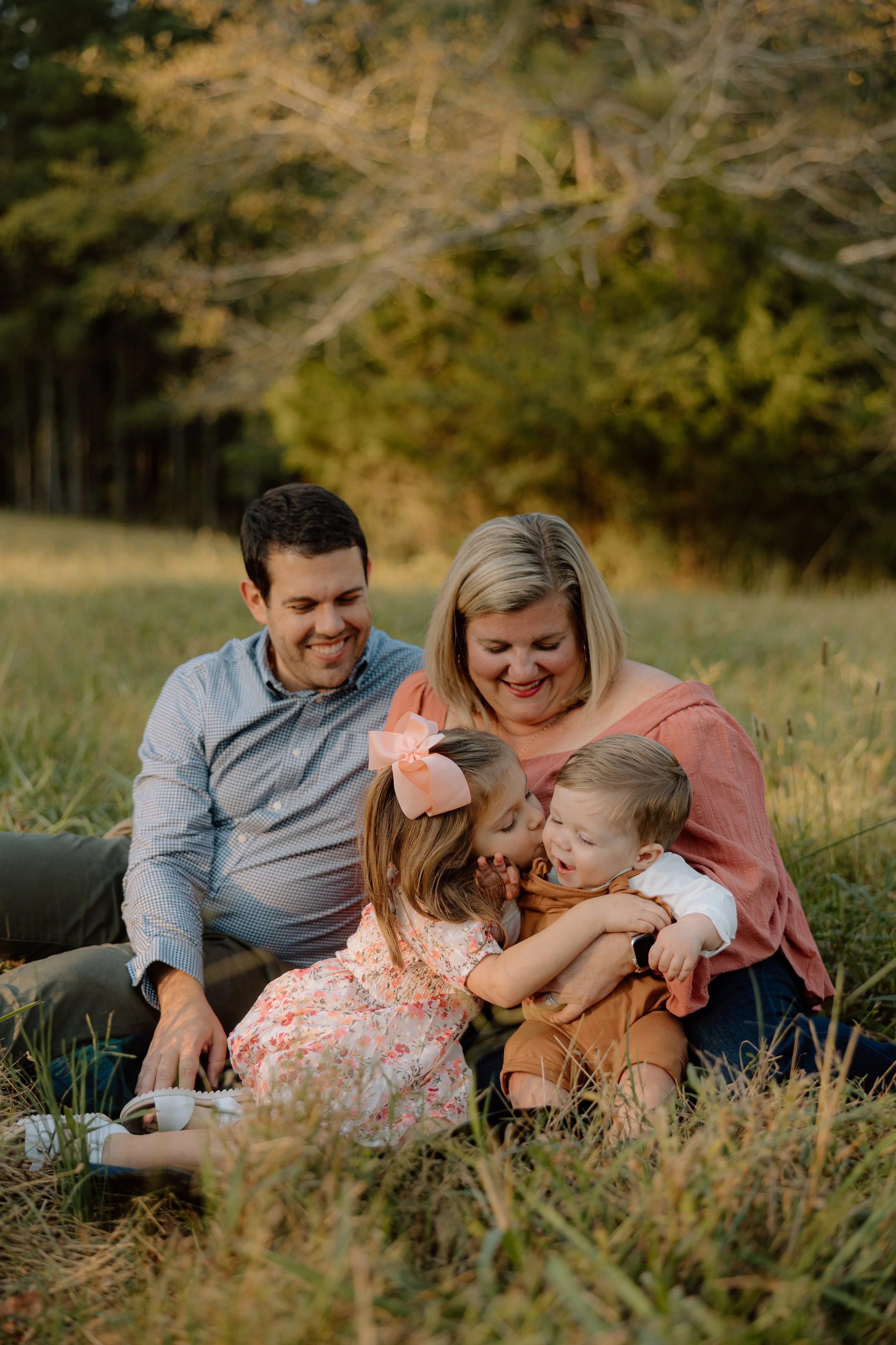 Family of four sitting on grass in a park, smiling and hugging, with trees and sunset in the background.