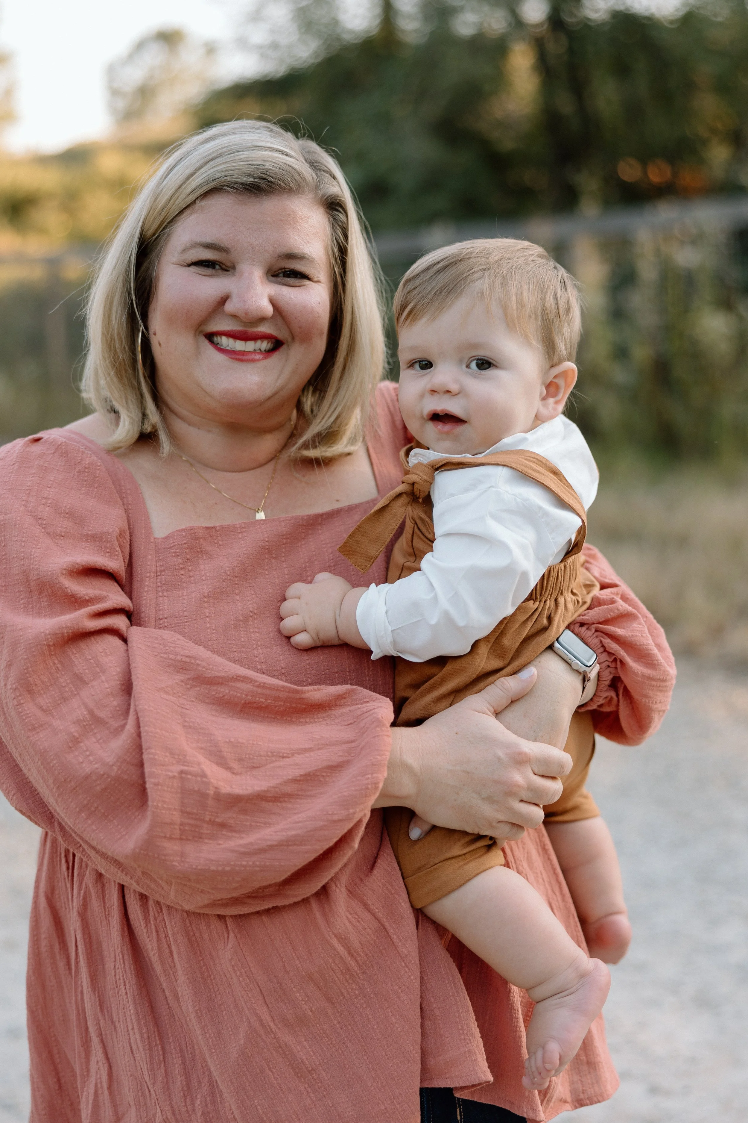 A woman with blonde hair smiling and holding a young boy outdoors near a body of water and trees in the background.