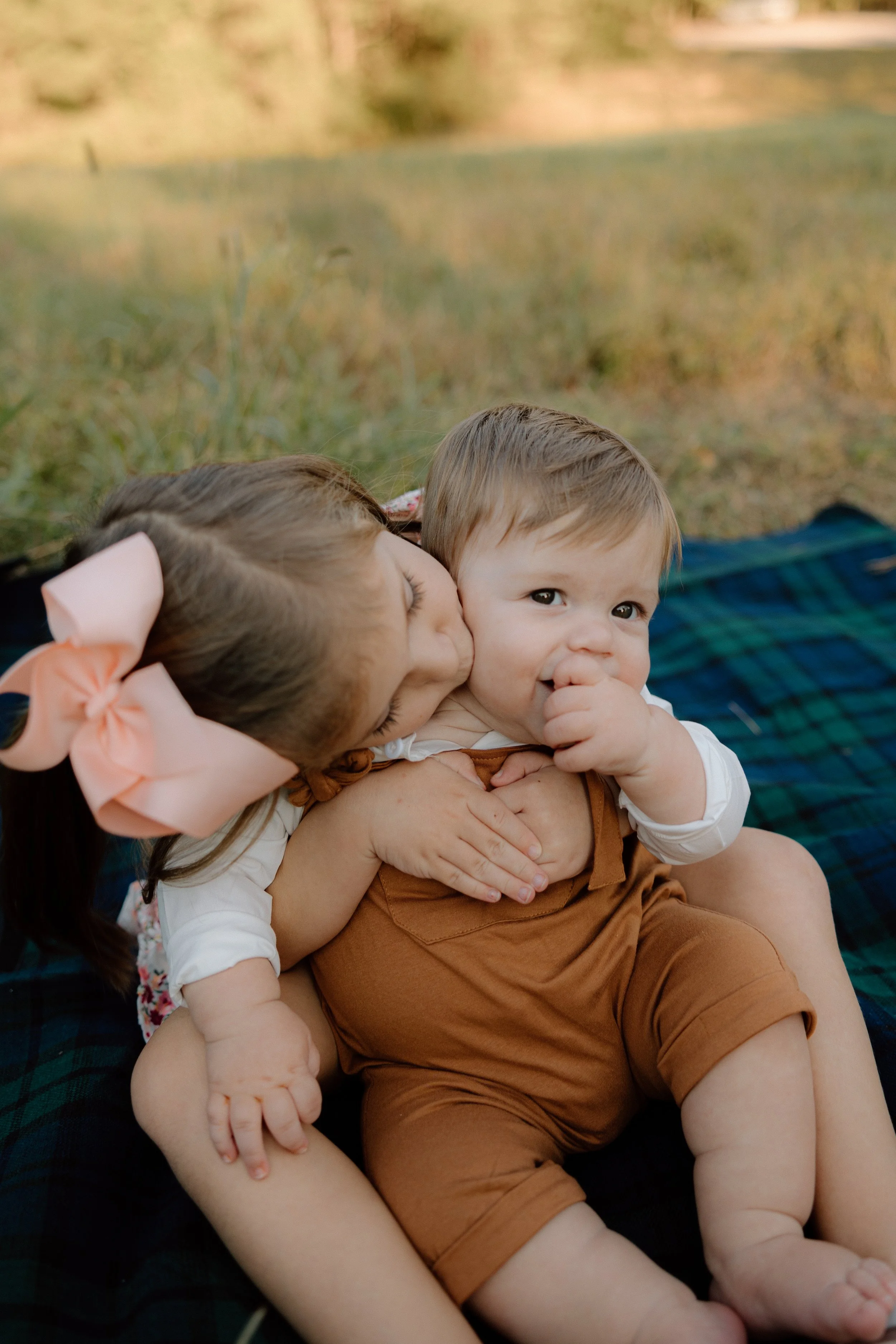 Two young children, a girl with a pink bow and a boy, sitting on a blanket outdoors. The girl is kissing the boy on the cheek while he smiles and looks at the camera.
