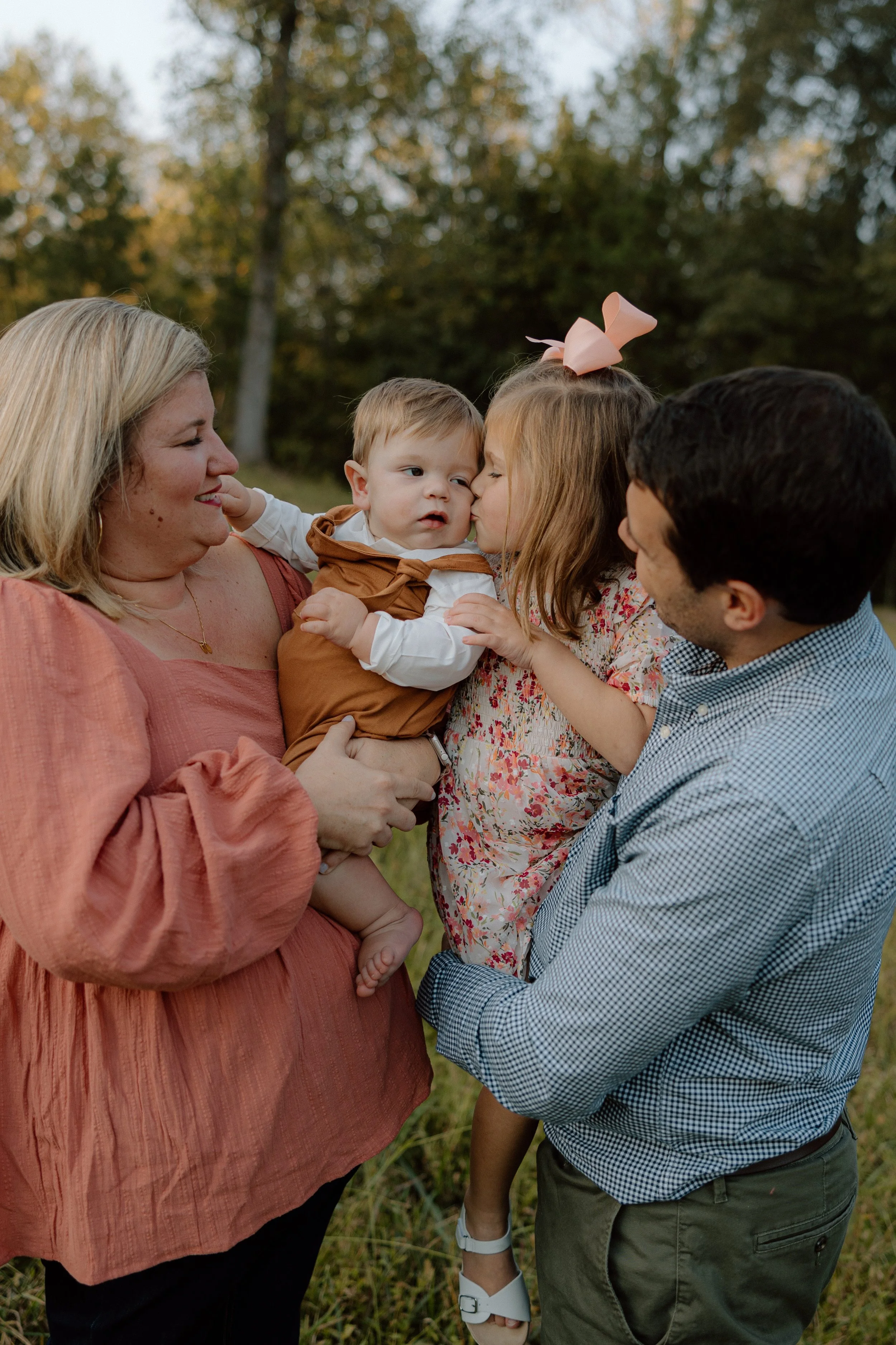 A family of four outside, with trees in the background, celebrating a moment together. The mother, with blonde hair, is holding a young boy in a brown outfit. The little girl is giving the boy a kiss, and the father is holding the girl. All are smili