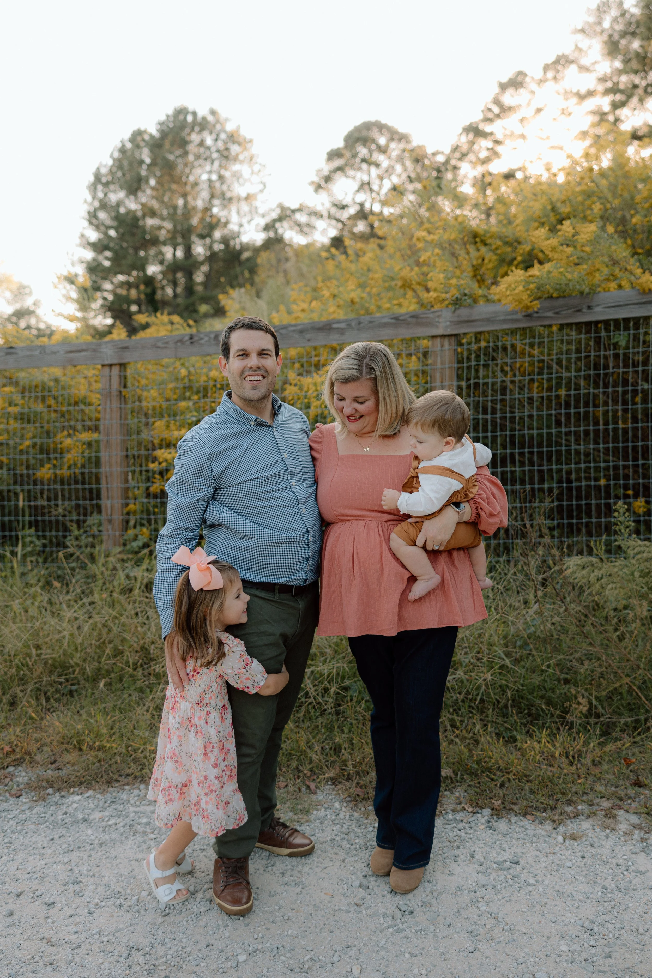 A family of four standing outdoors in front of a wooden fence and yellow fall foliage, smiling and enjoying the moment.