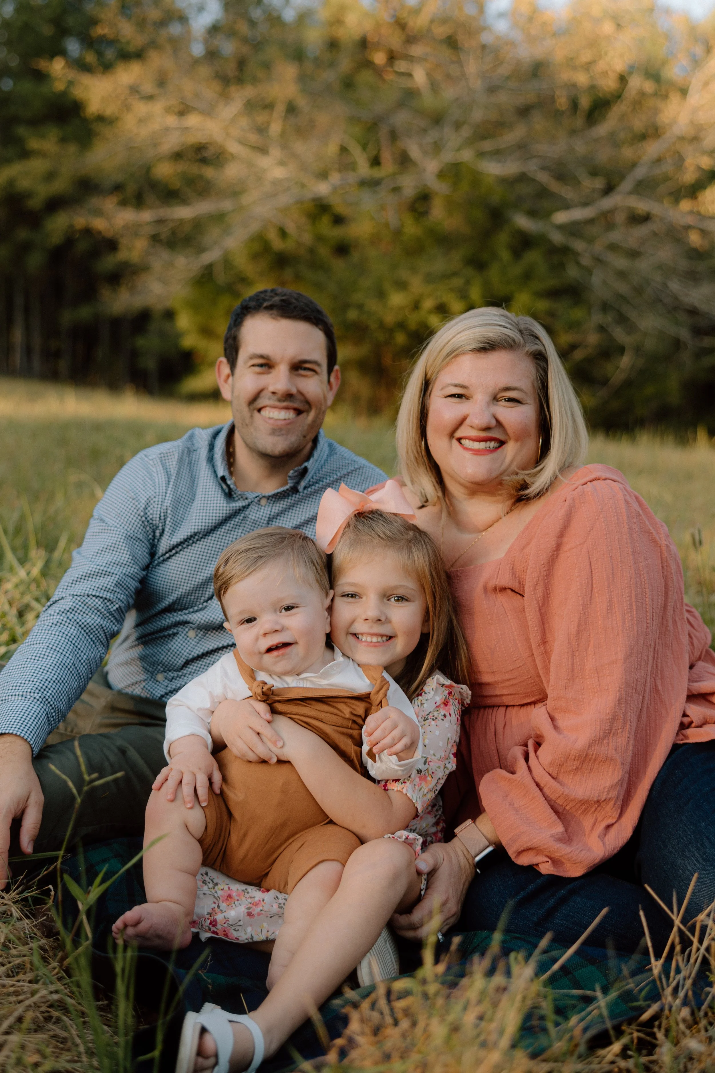 A happy family of four sitting on the grass in a park during late afternoon, smiling at the camera, with trees in the background.