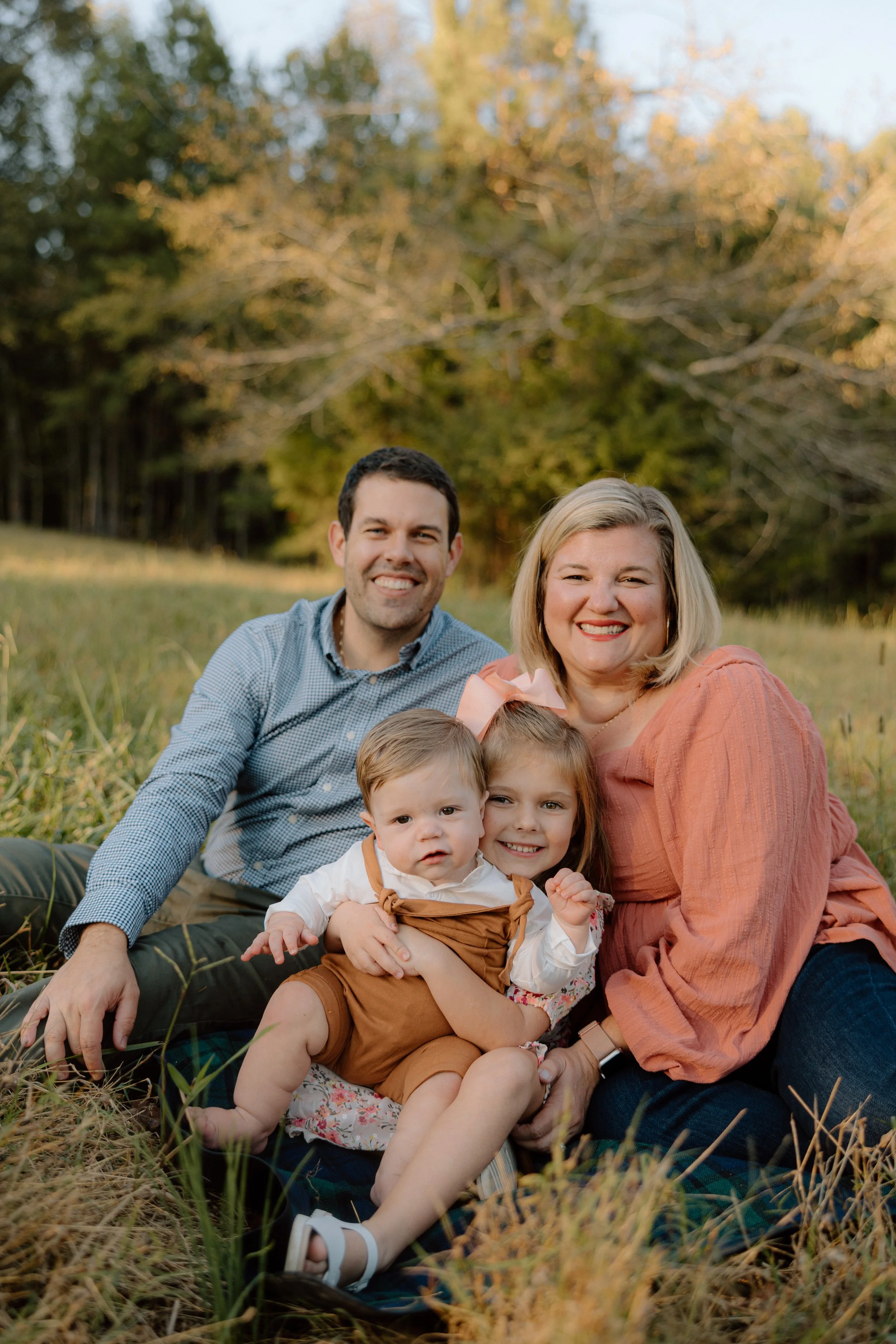 Family of four sitting on grass in a field with trees in the background, smiling.