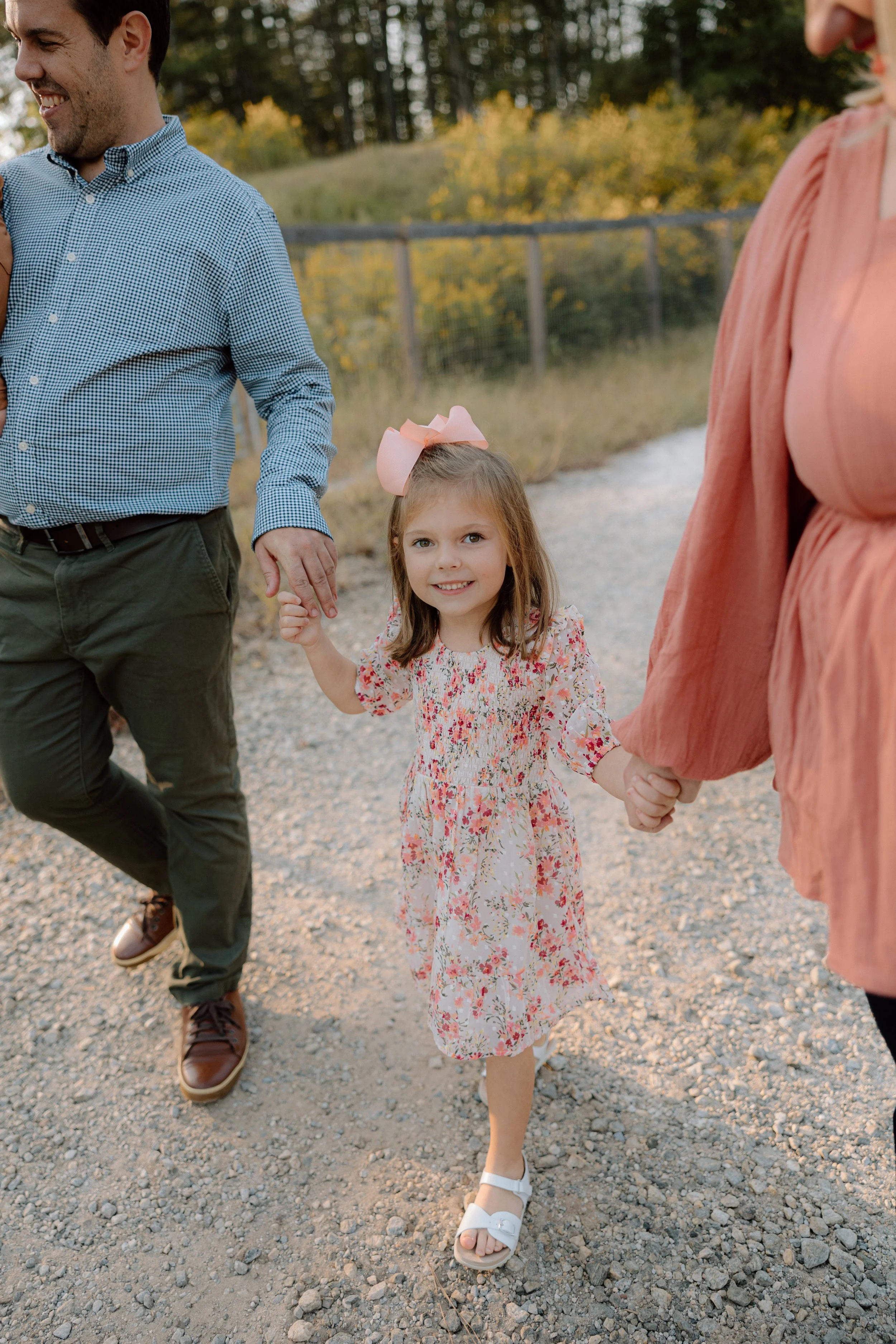 A young girl with a pink bow in her hair, wearing a floral dress and white sandals, is holding hands with two adults, a man and a woman, as they walk outdoors on a gravel path during the daytime.