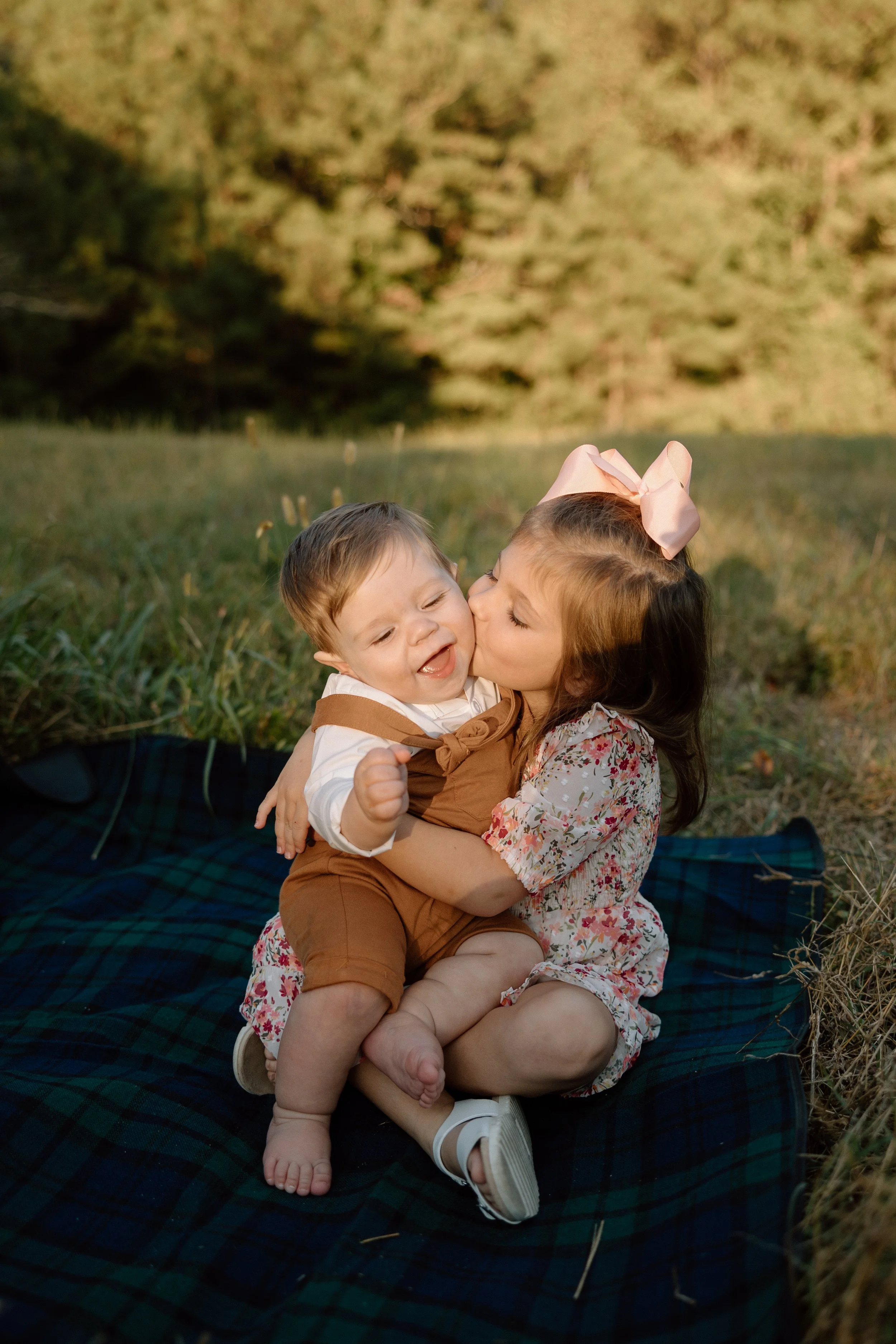 A young girl and boy sitting on a blanket outdoors, with the girl kissing the boy on the cheek, in a grassy area with trees in the background.
