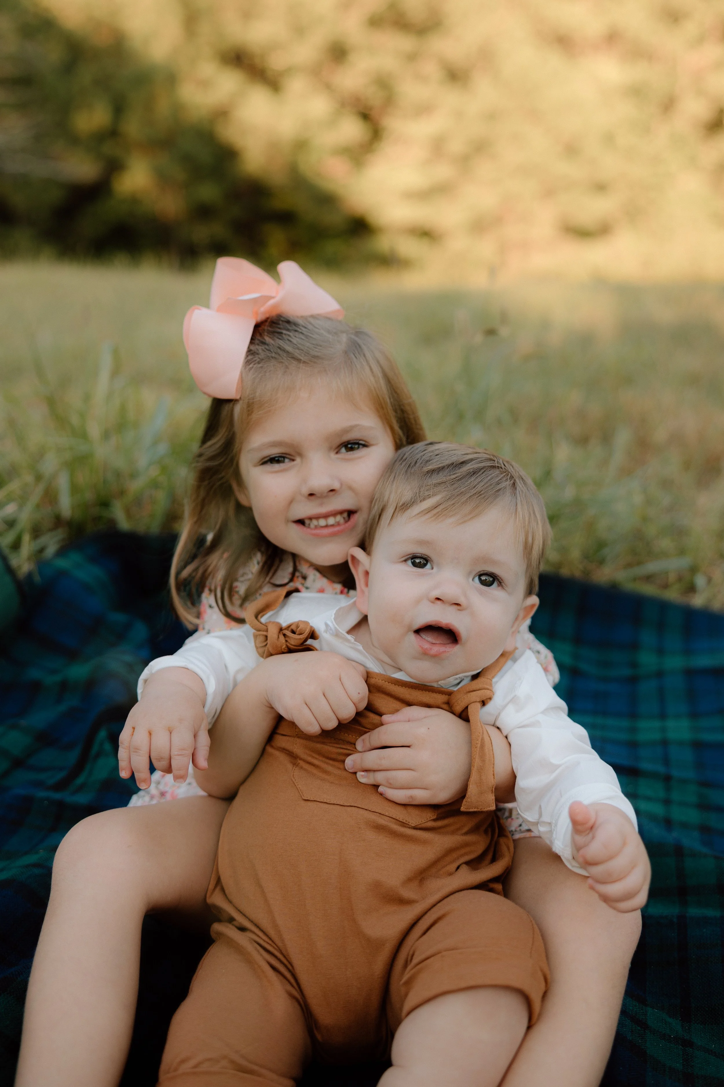 Two young children, a girl with a large pink bow in her hair and a boy with short light brown hair, sitting together on a blanket outdoors in a grassy area.