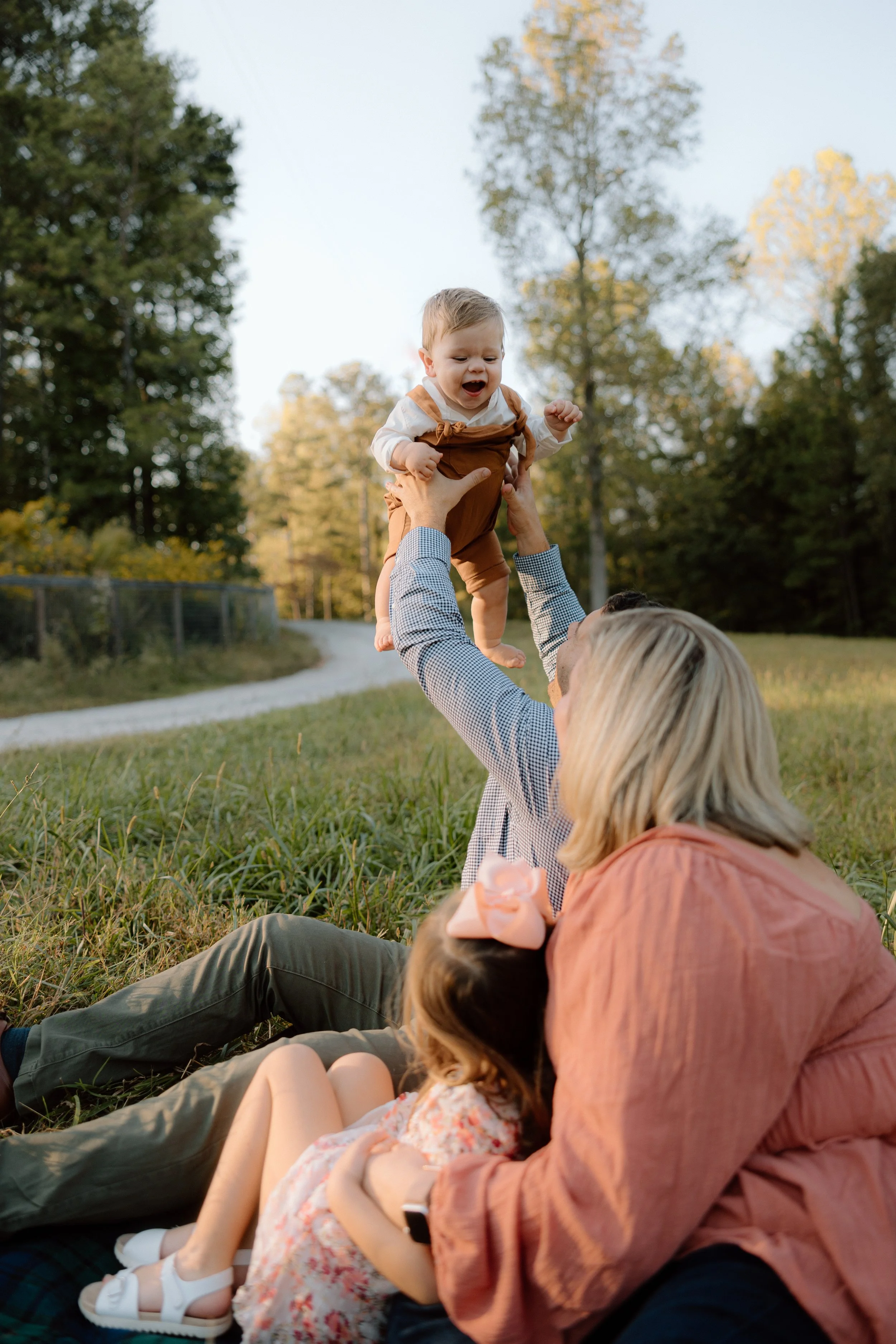 Family outdoors on a grassy field, with a woman and a man sitting on the ground, and a young girl with a pink bow sitting between them. The man is lifting a happy toddler boy into the air, and trees in the background.