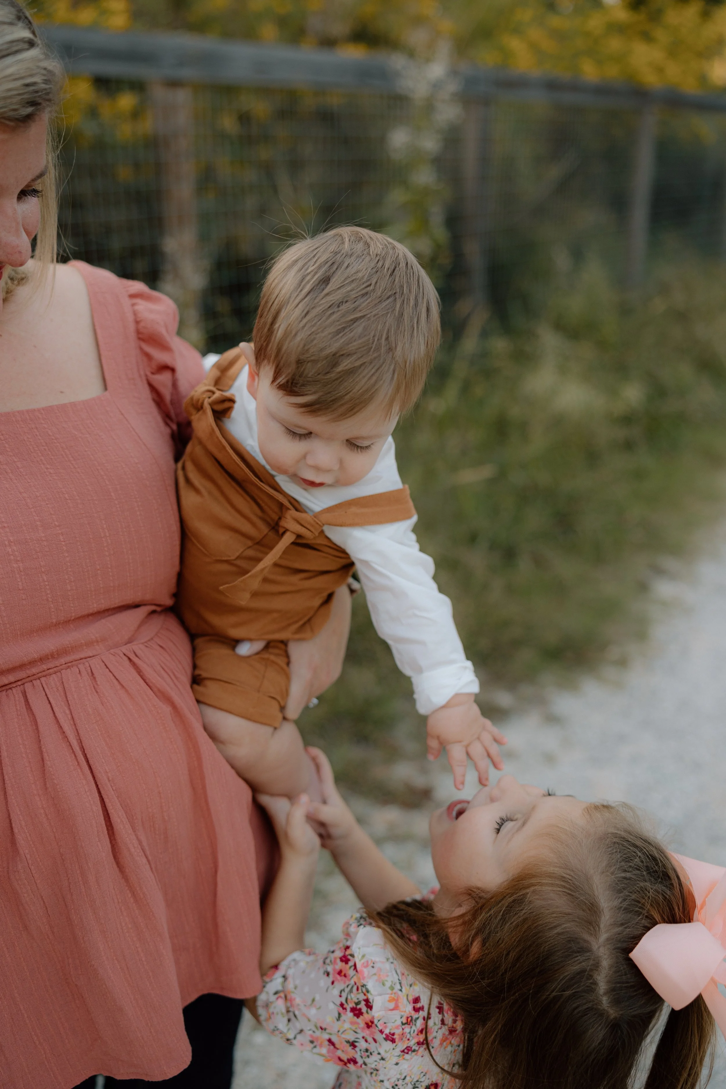A woman holding a young boy outside near a fence, with a girl reaching up towards the boy's face, all smiling and playing together.