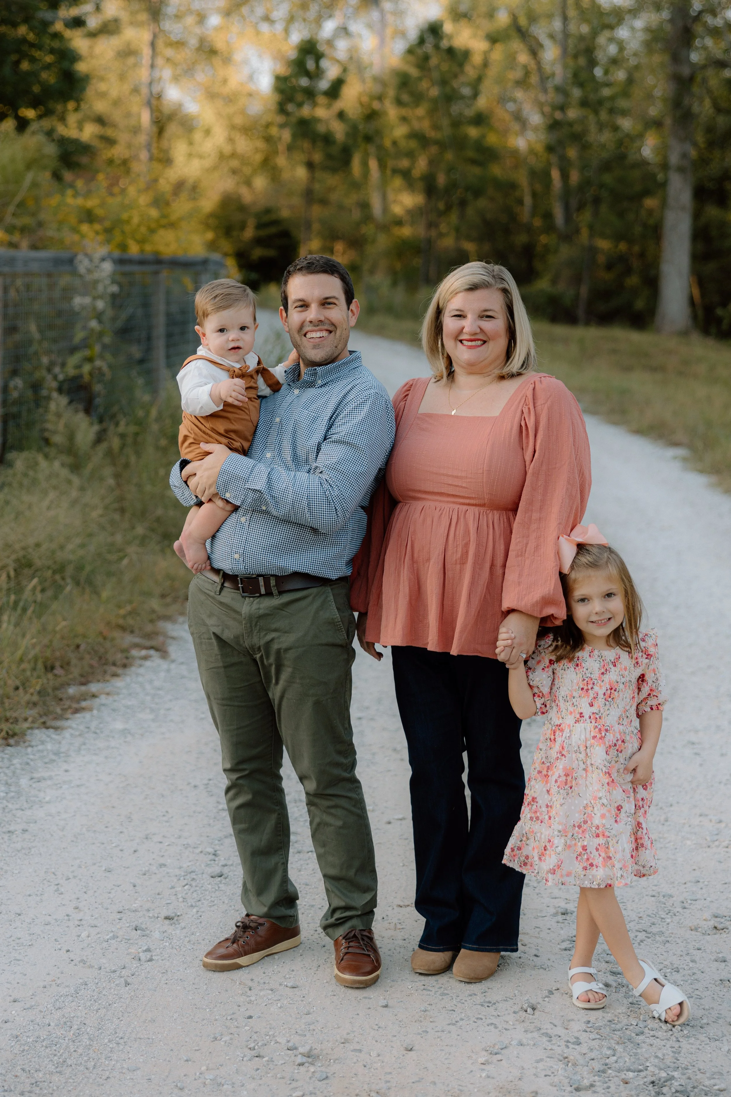 A family of four standing on a gravel path outdoors, smiling at the camera with trees in the background. The father is holding a young child, and the mother is holding the hand of an older girl.