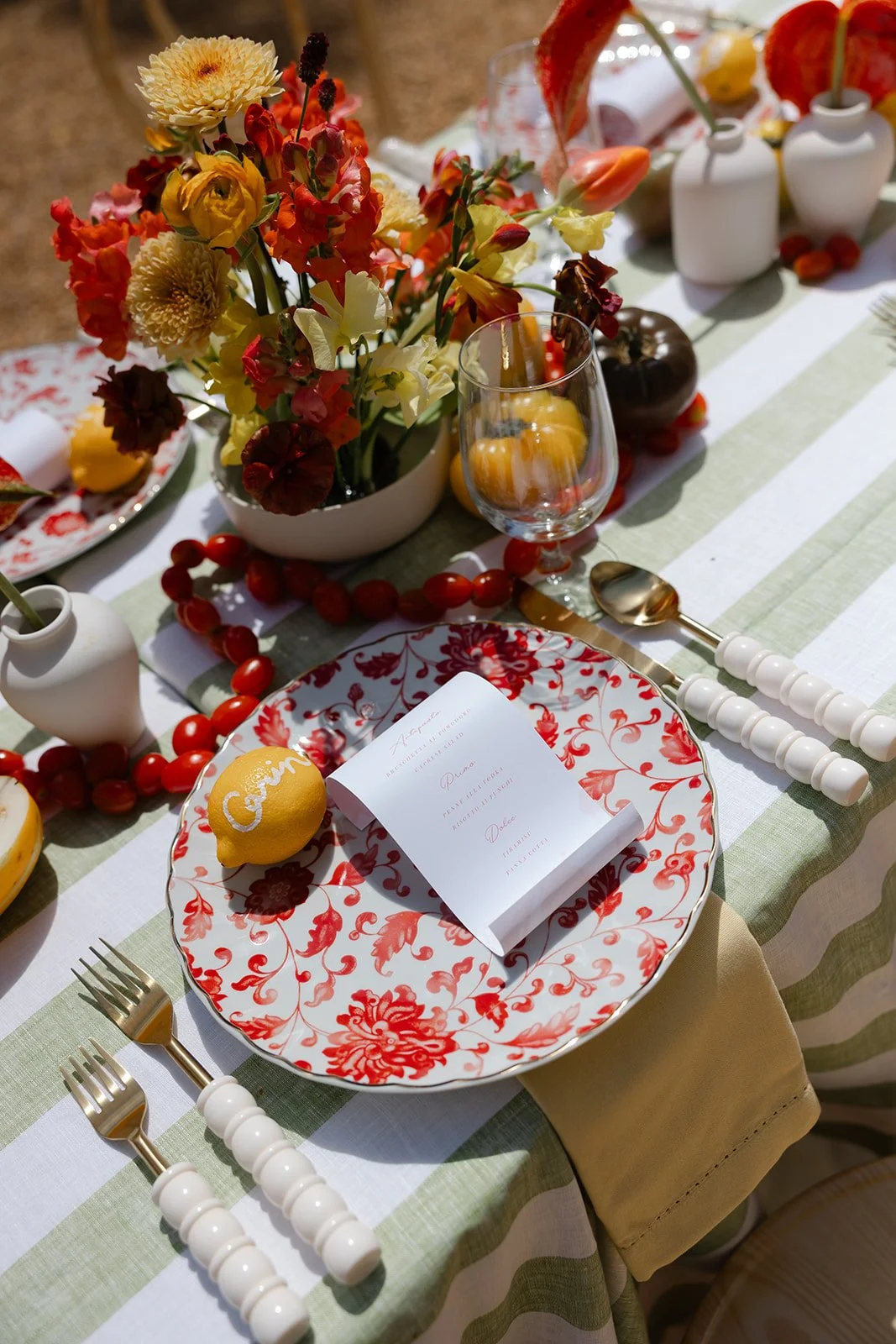 A table setting with a red and white floral plate, a lemon with white writing, cutlery with white beaded handles, a floral centerpiece, and a printed menu on the plate.