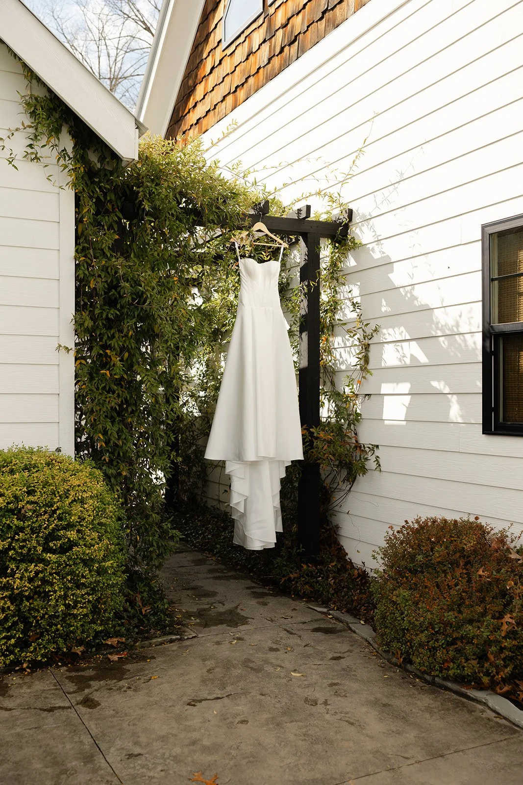 A white wedding dress hanging on a black wooden trellis between white house siding and a bush, with shadows cast on the wall and a window nearby.