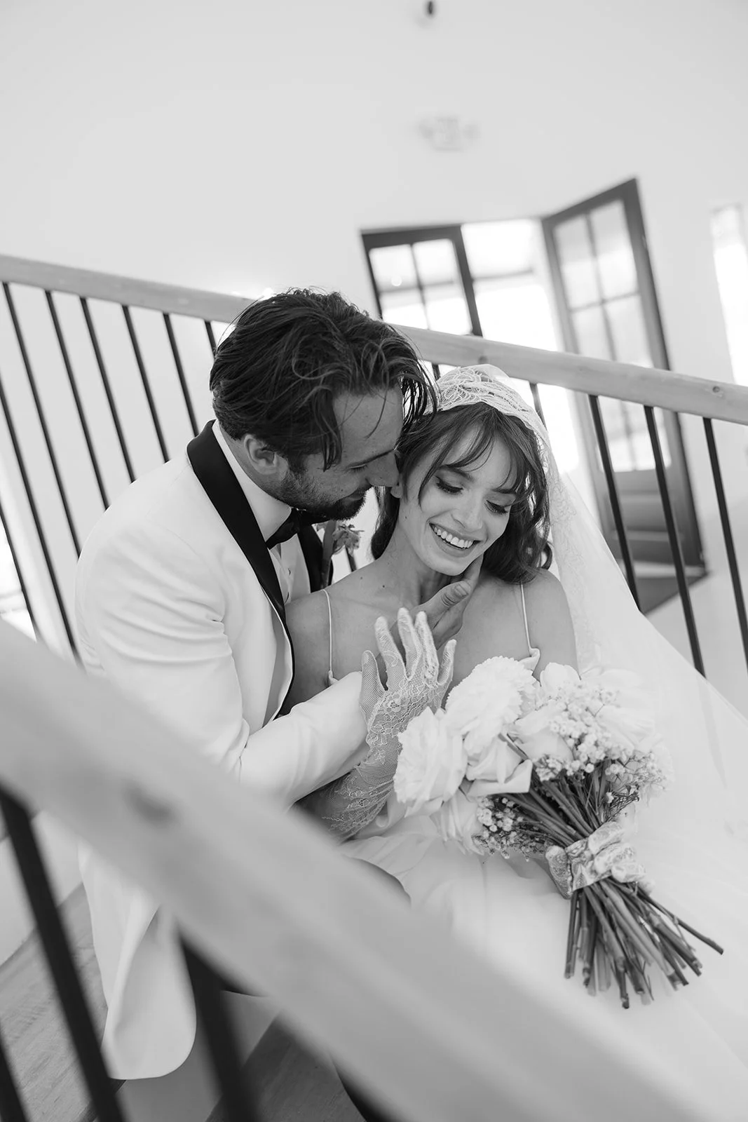 A black-and-white photo of a bride and groom sitting on stairs. The groom is dressed in a tuxedo, leaning close and touching the bride's face. The bride is wearing a wedding gown, holding a bouquet and smiling joyfully. She has a veil and lace gloves