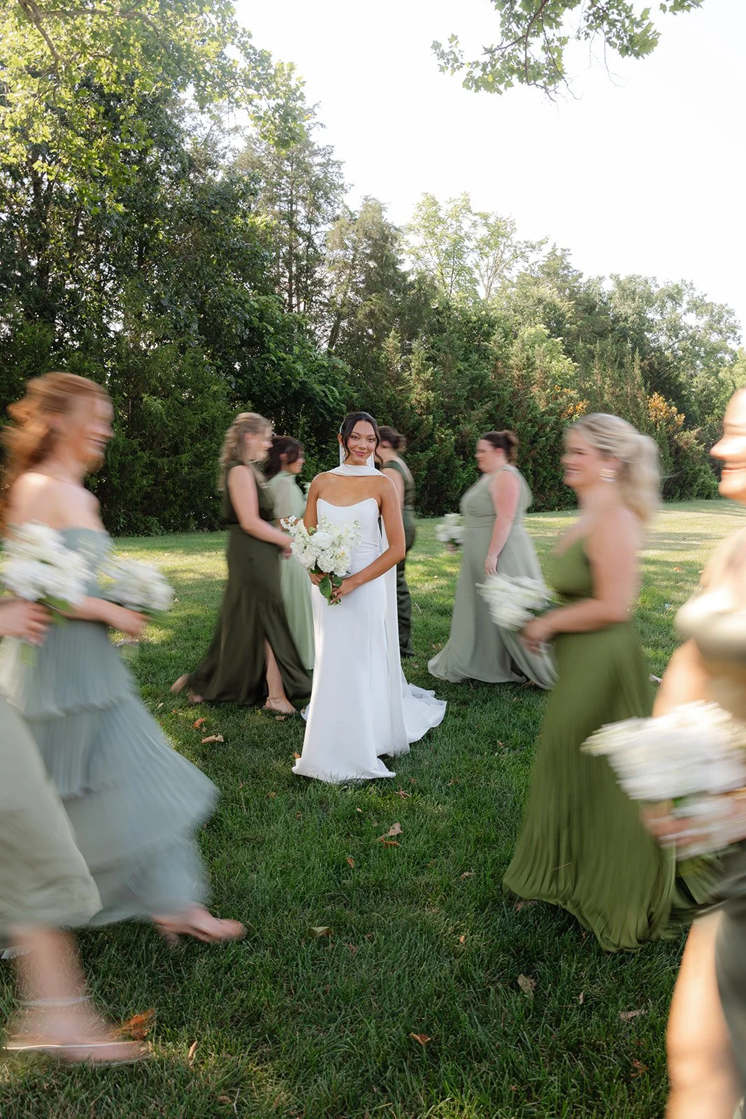 A bride in a white wedding dress holding a bouquet of white flowers standing in a grassy outdoor setting, surrounded by bridesmaids in shades of green and gray dresses holding similar bouquets, with a background of trees and a bright sky.