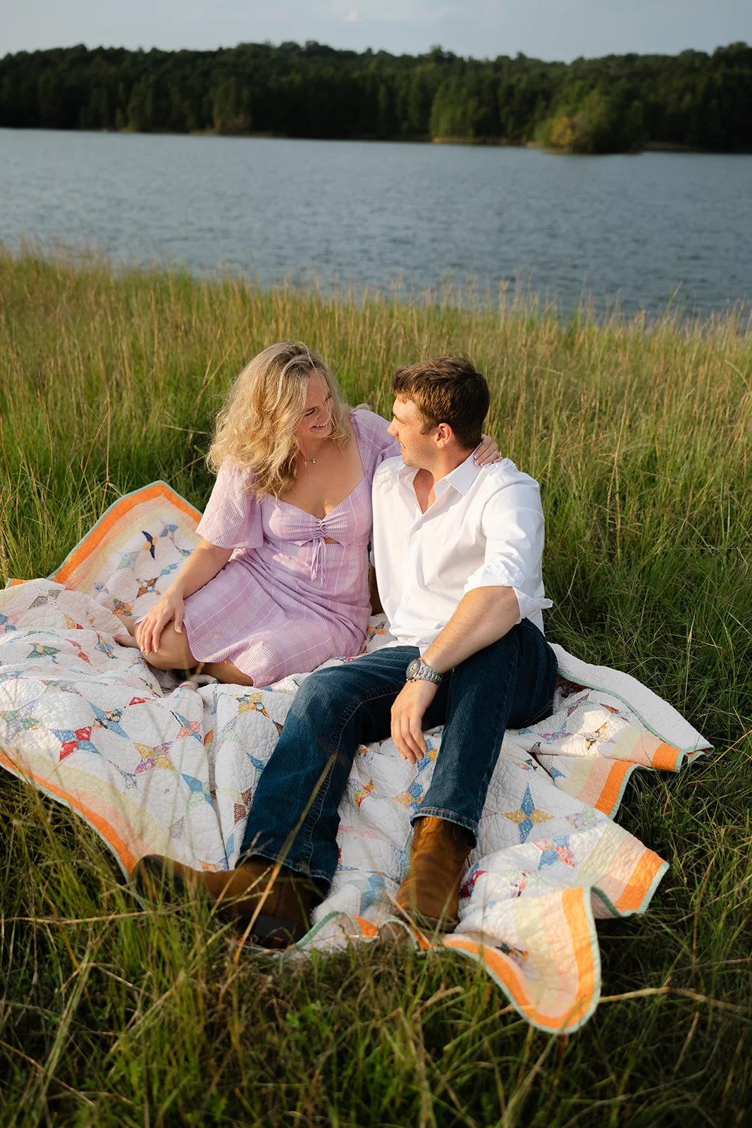 A couple sitting on a quilt by a lake, surrounded by grass and trees, enjoying a sunny day.