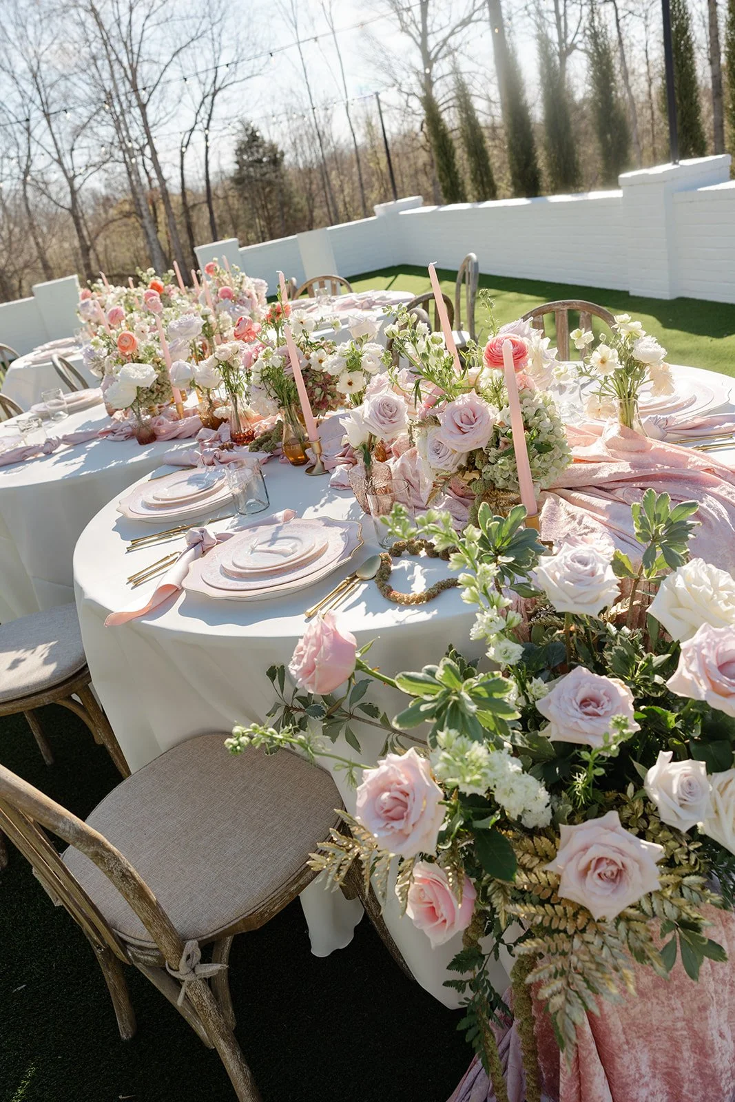 Outdoor wedding reception table with pink and white floral arrangements, pink candles, and elegant tableware.