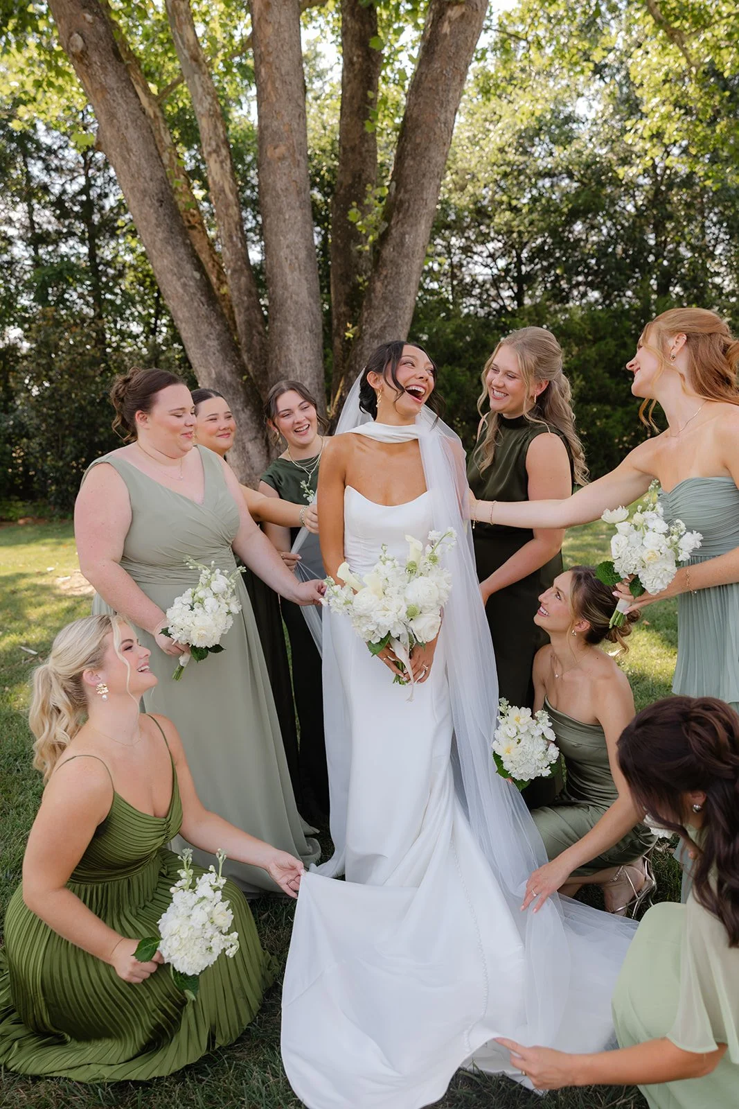 A bride surrounded by her bridesmaids outdoors, laughing and smiling, holding white bouquets, with a large tree in the background.